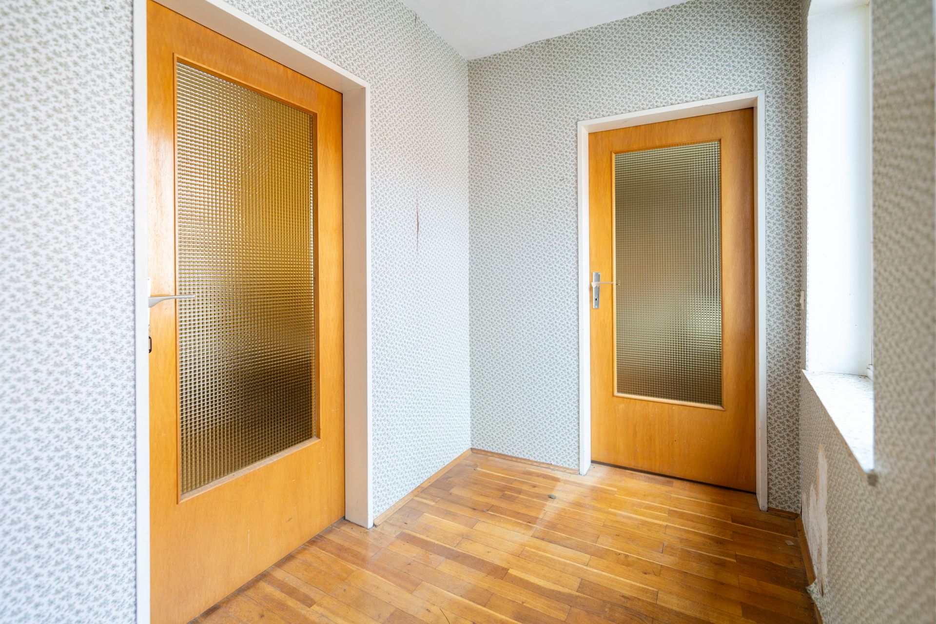 Heller Flur mit zwei Holztüren und einem Fenster, Parkettboden und gemusterter Tapete.