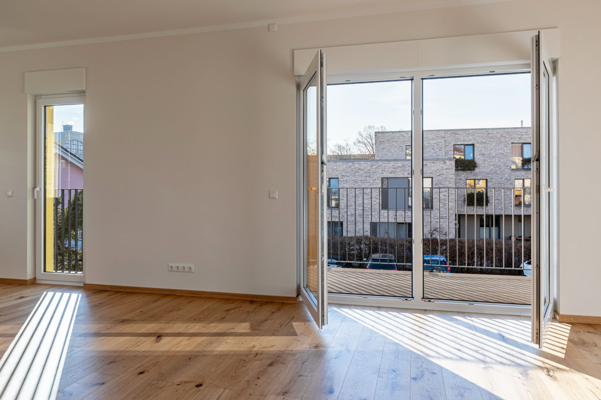 Leerer Raum mit Holzboden, geöffneten Glastüren zum Balkon und Blick auf moderne Wohngebäude.