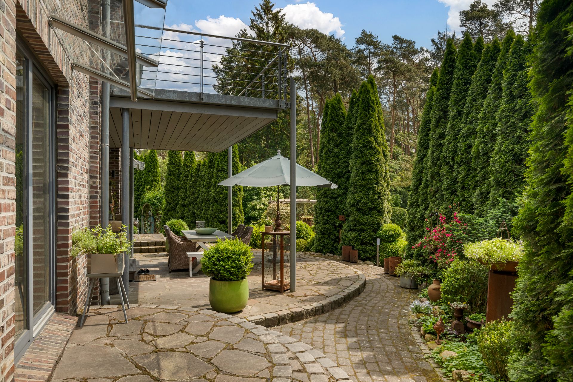 Steinterrasse mit Sitzbereich, Sonnenschirm und vielen grünen Pflanzen neben einem Wohnhaus.