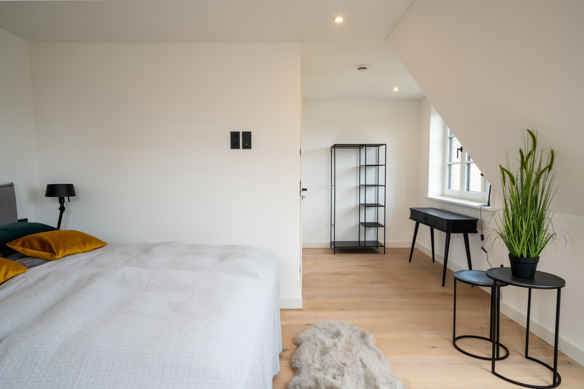 Helles Schlafzimmer mit Bett, Schreibtisch, Regal und Pflanze am Fenster.