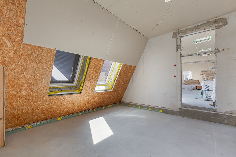 Zimmer im Rohbau mit schräger Decke, zwei Dachfenstern und offenem Durchgang zu weiterem Raum.