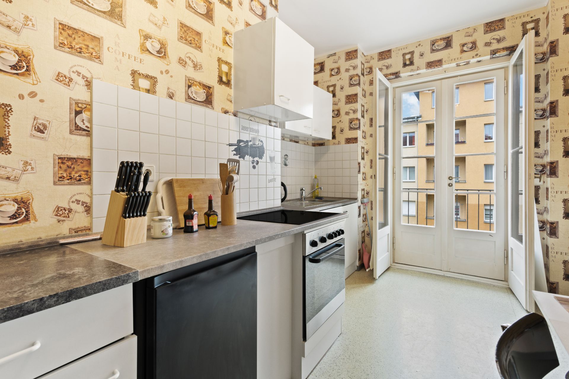 Kleine Küche mit Kaffeemotiv-Tapete, Herd, Spüle, Messerblock und Türen zum Balkon.