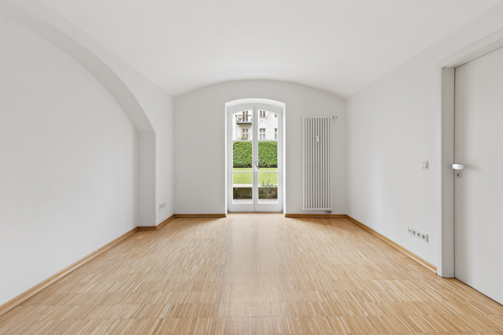 Heller, leerer Raum mit Holzboden, weißem Heizkörper und Glastür zum Garten.