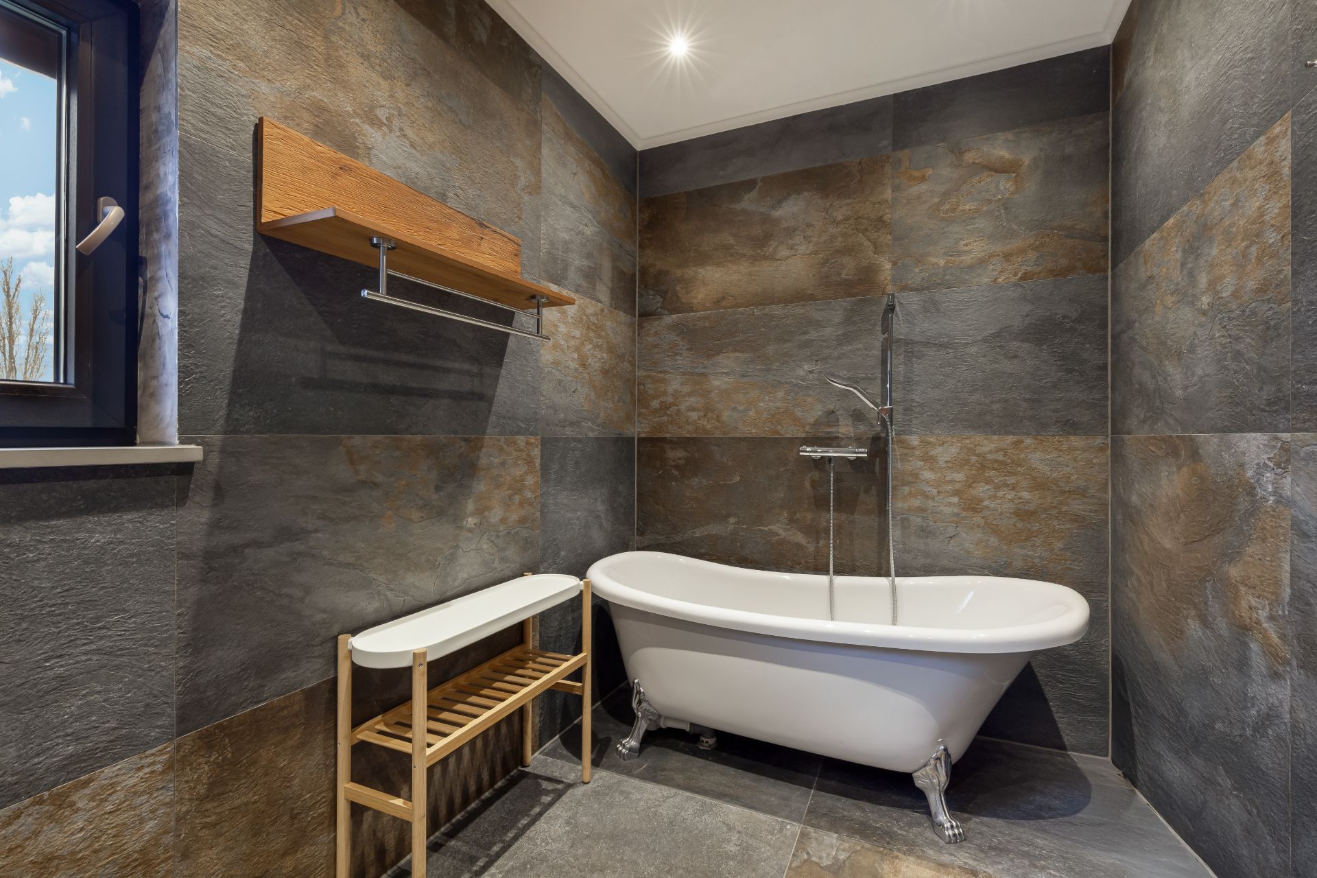 Modernes Badezimmer mit freistehender Badewanne, dunklen Fliesen und Holzregal an der Wand.