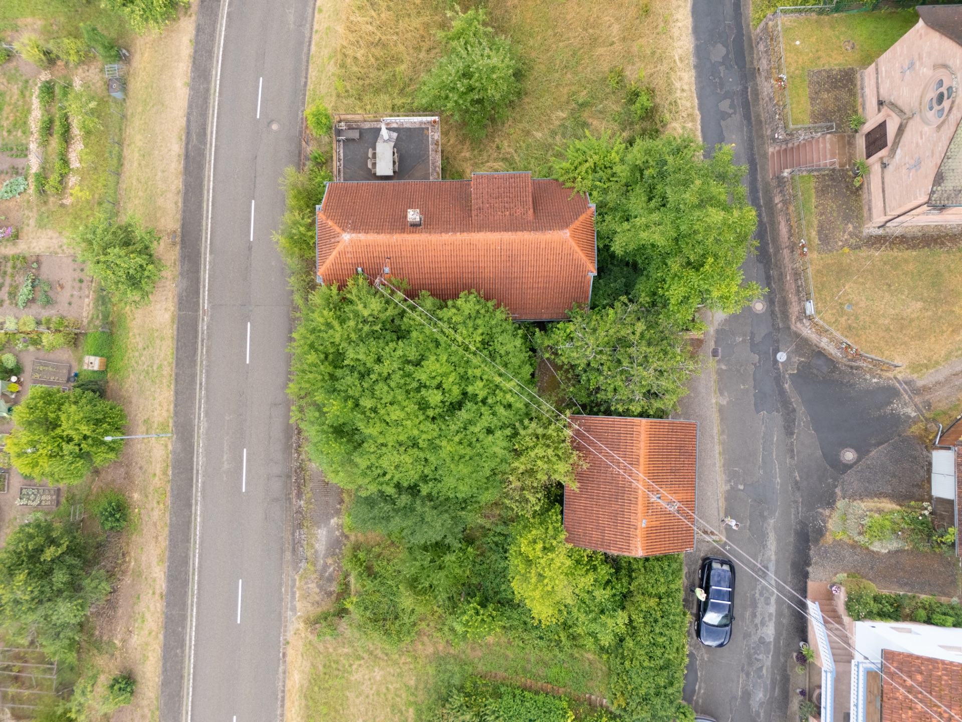 Luftaufnahme eines Hauses mit rotem Dach, umgeben von Bäumen, an einer Straßenecke mit geparktem Auto.