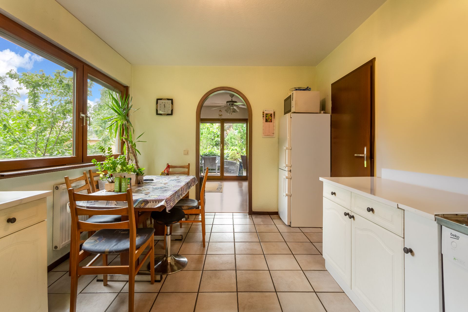 Helle Küche mit großem Fenster, Esstisch, Stühlen, Kühlschrank und Pflanzen.