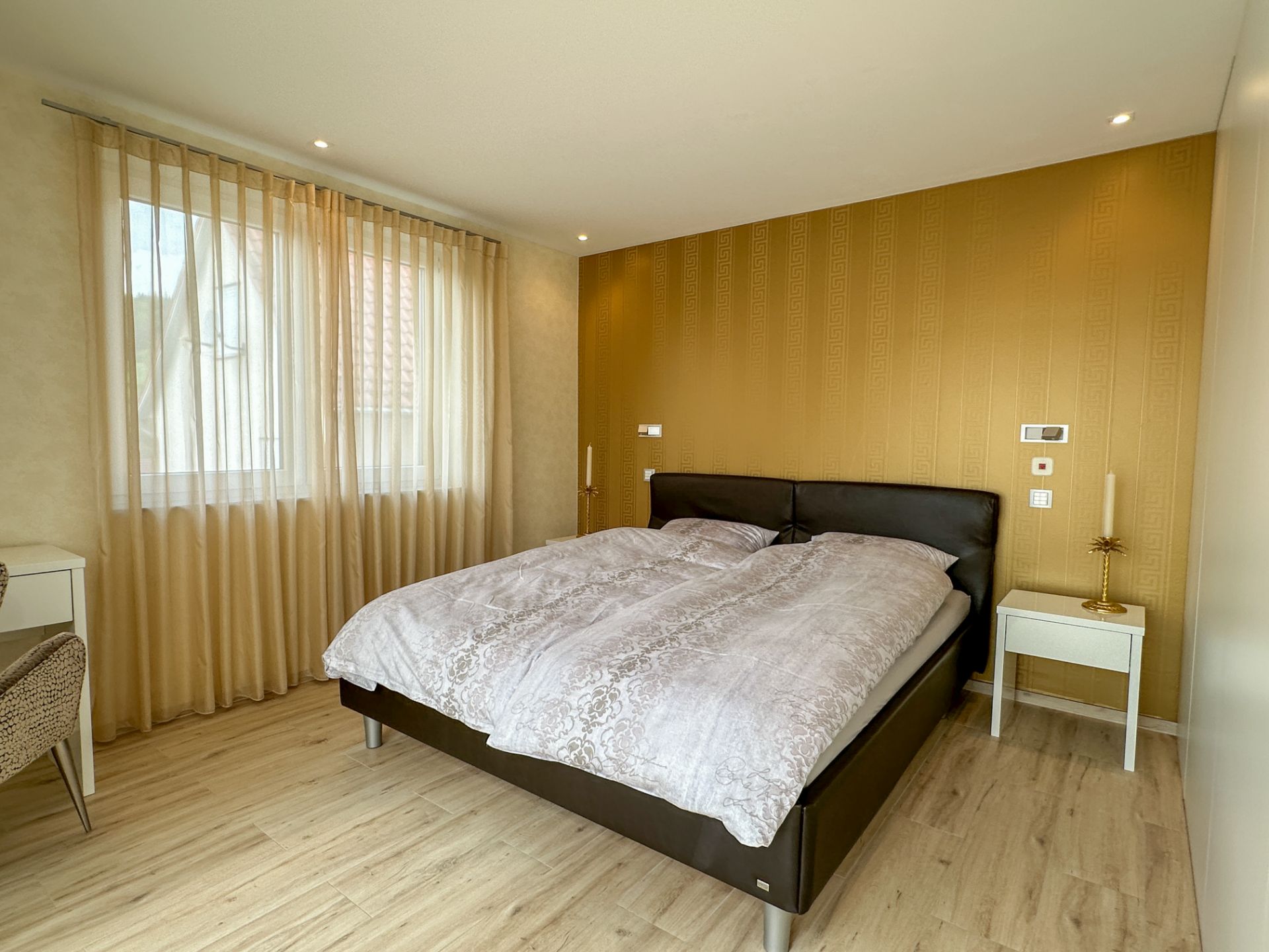 Modernes Schlafzimmer mit Doppelbett, goldener Akzentwand und großem Fenster mit Vorhängen.