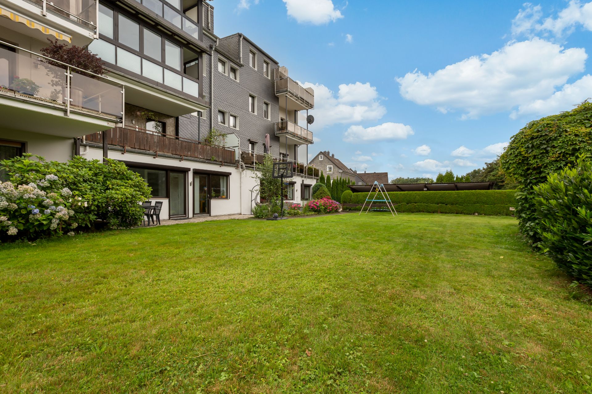 Grüner Garten mit Rasenfläche, Schaukel und mehrstöckigem Wohngebäude im Hintergrund.