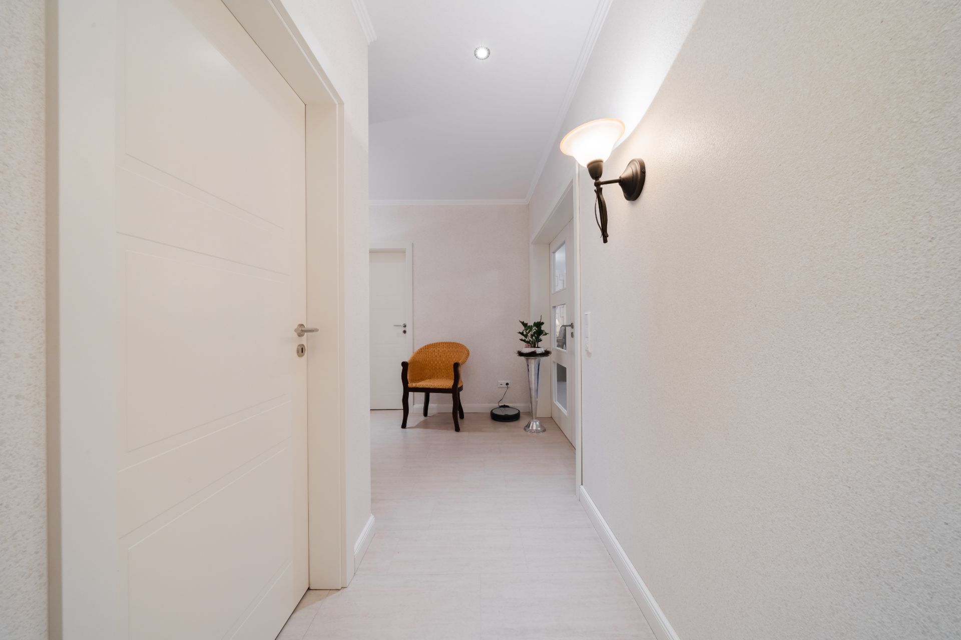 Heller Flur mit weißer Tür, Wandlampe, Stuhl und Pflanze auf einem kleinen Tisch.