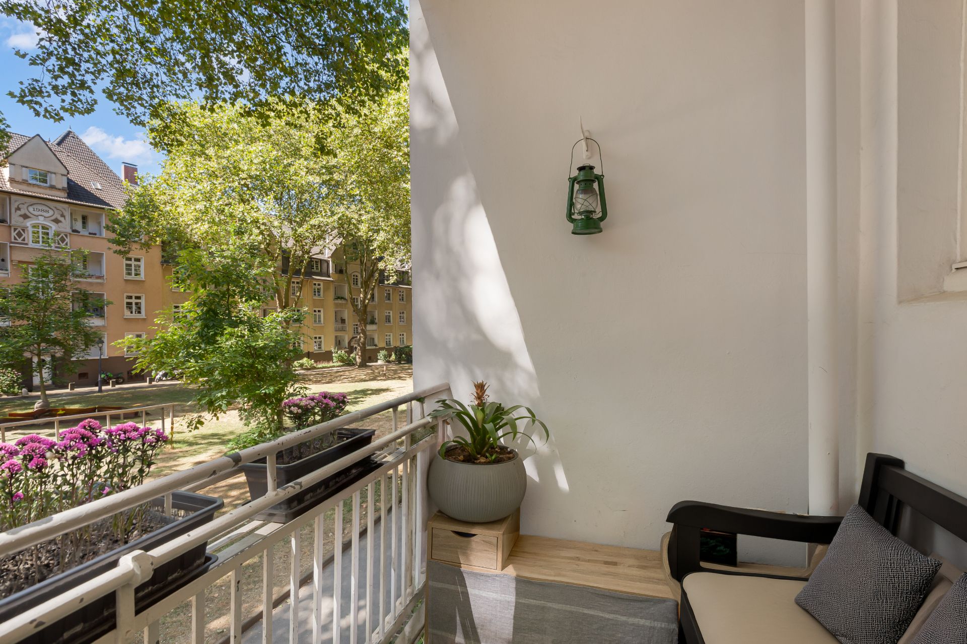 Balkon mit Bank, Pflanze und Laterne, Blick auf Garten und Wohnhäuser im Hintergrund.