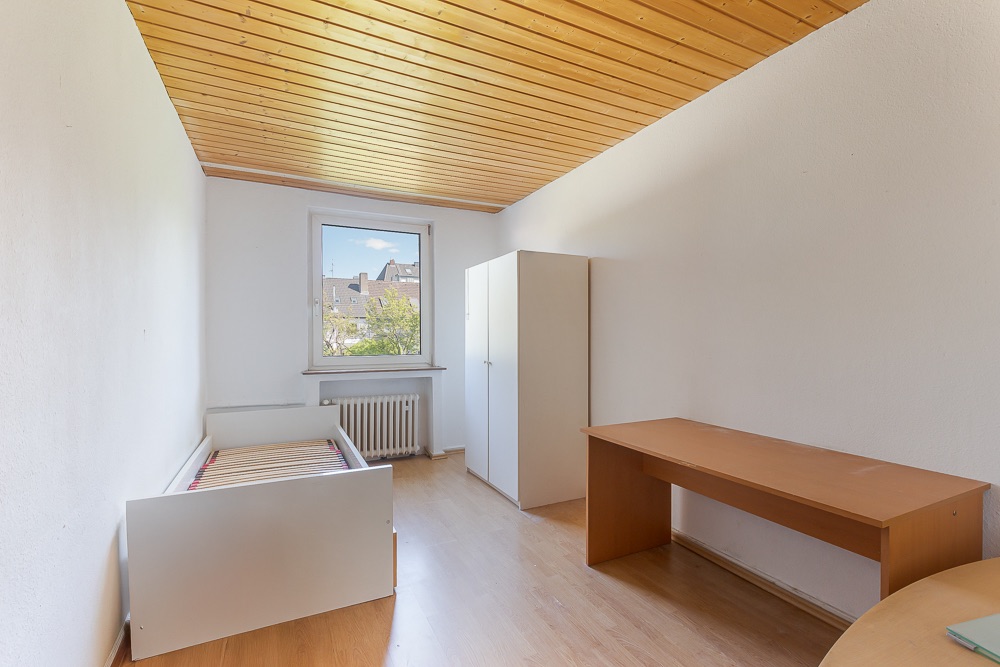Schlichtes Zimmer mit Einzelbett, Schreibtisch, Kleiderschrank und Fenster mit Blick nach draußen.