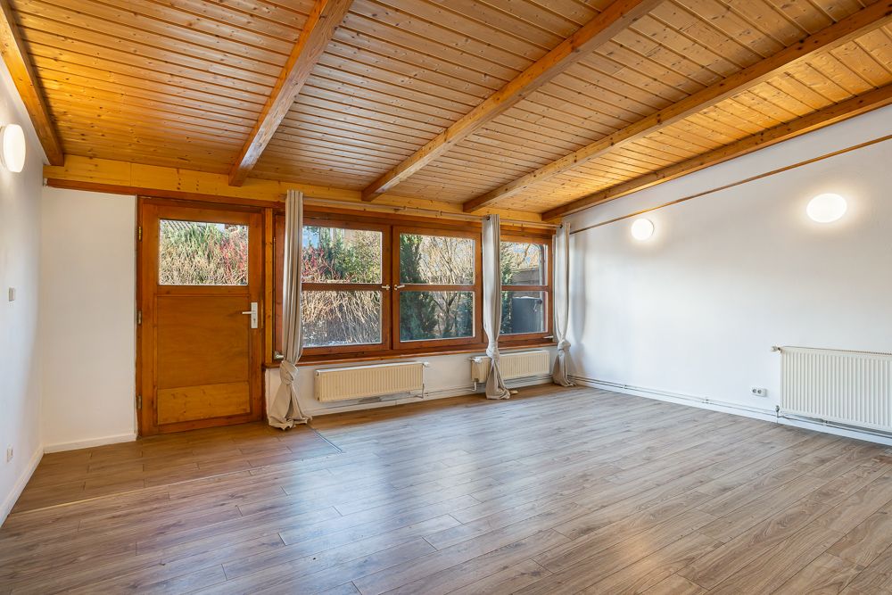 Helles Wohnzimmer mit Holzdecke, großem Fenster, Holztür und Heizkörpern, leer und mit Vorhängen.
