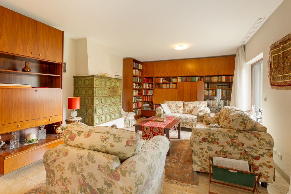 Wohnzimmer mit geblümten Sofas, grünem Kachelofen, Bücherregalen und Holzmöbeln.