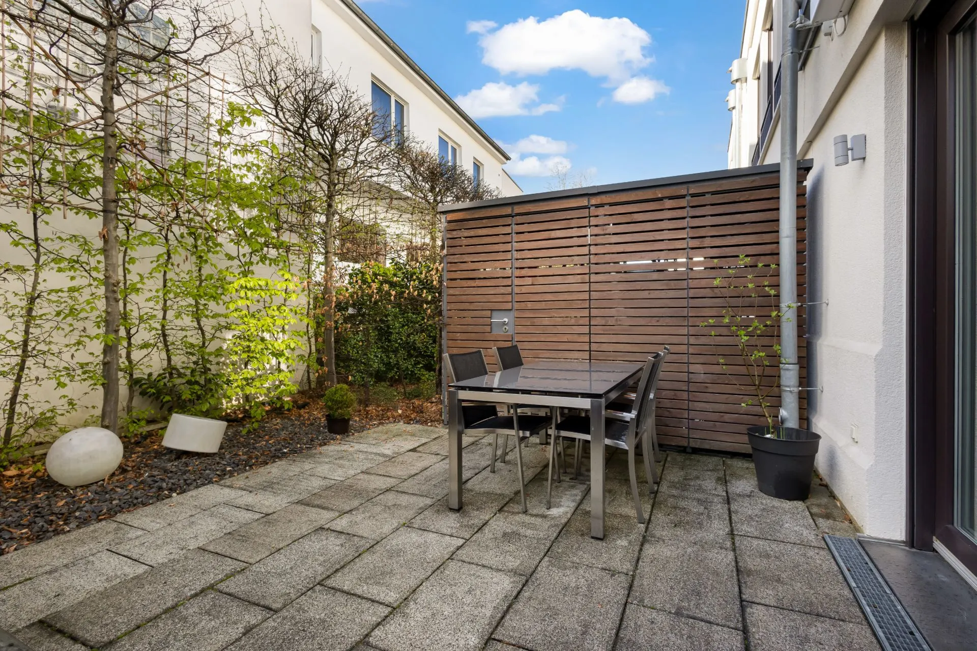 Gepflasterte Terrasse mit Tisch, vier Stühlen und Pflanzen, von Holzwand und Büschen umgeben.