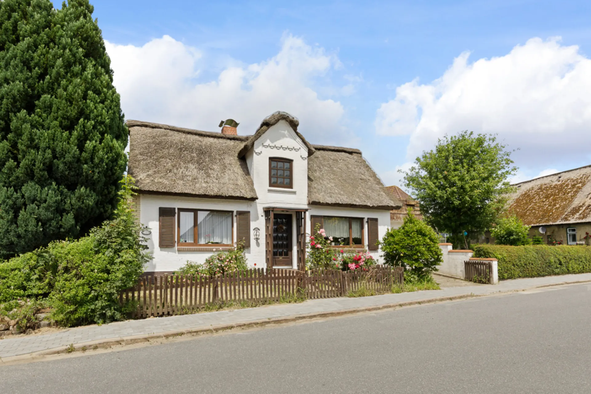 Weißes Wohnhaus mit Reetdach, Vorgarten, Holzzaun und grünen Pflanzen an einer Straße.