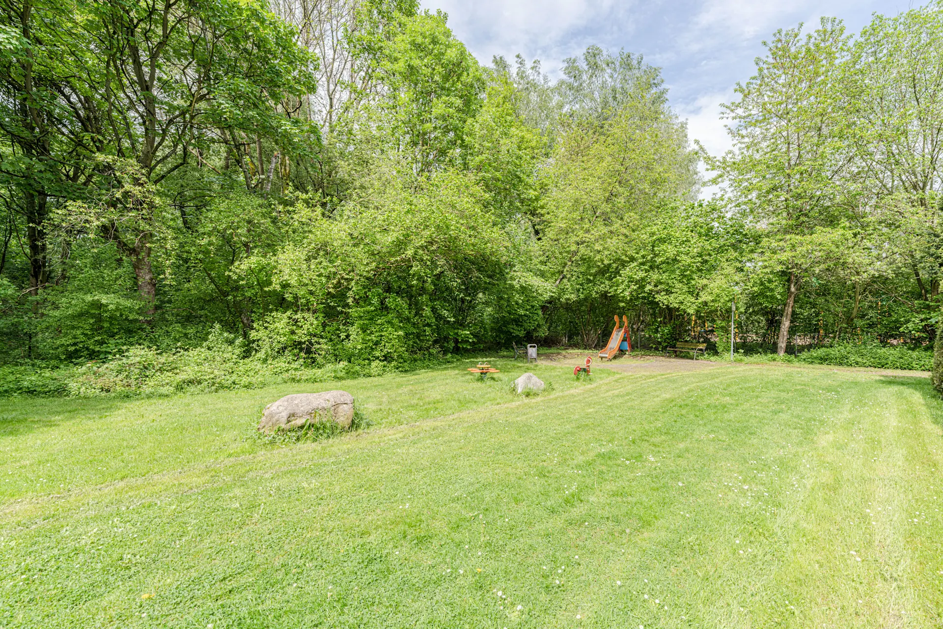 Grüner Garten mit Rasen, Bäumen, Felsen und Kinderspielplatz mit Rutsche.