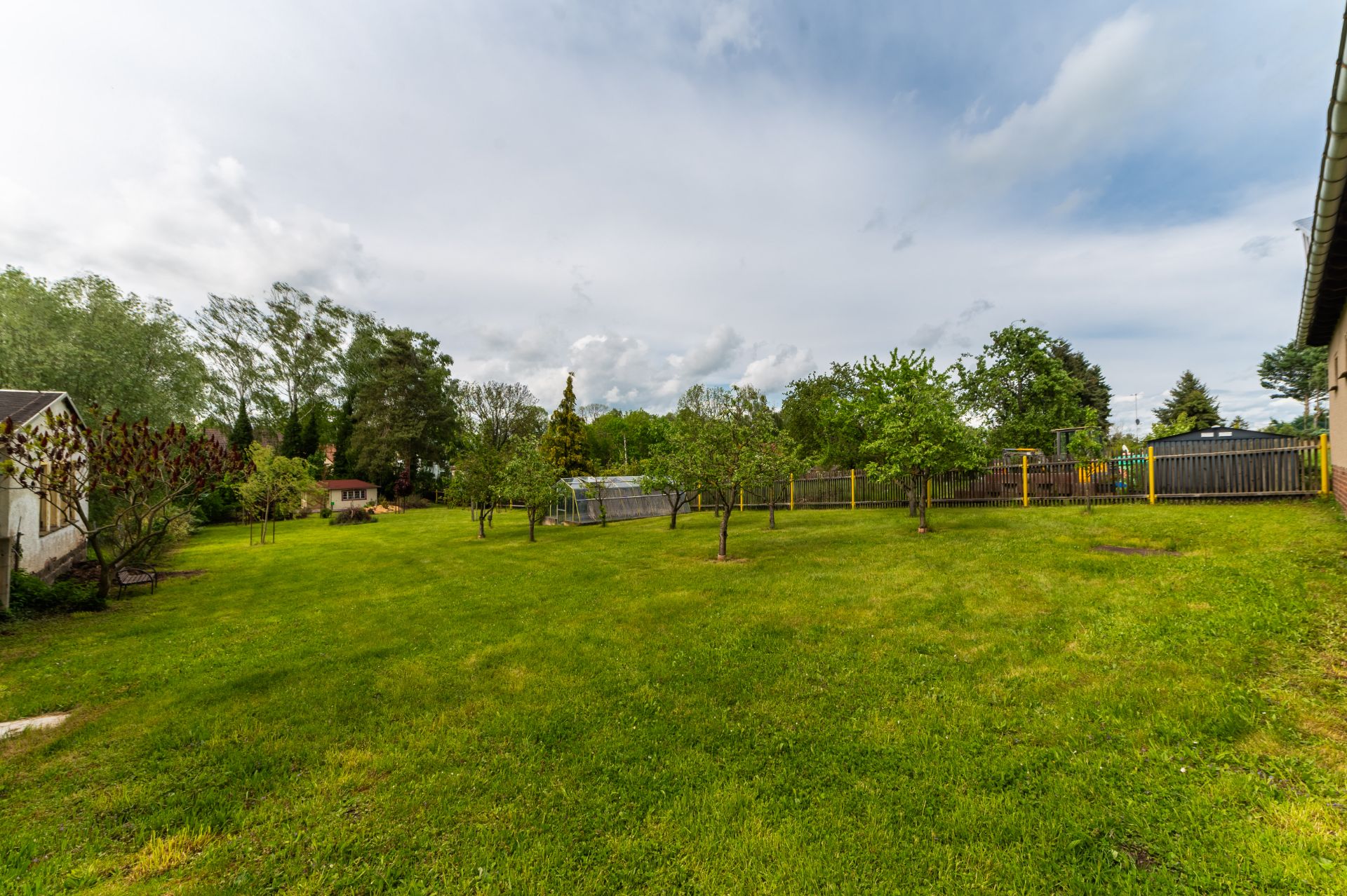 Großer grüner Garten mit Bäumen und umzäunten Bereich, Häuser im Hintergrund.
