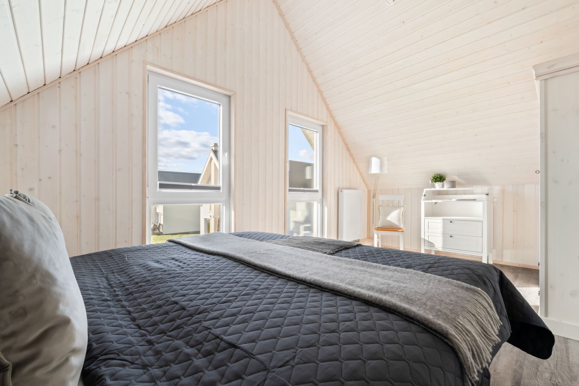 Helles Schlafzimmer mit großem Bett, zwei Fenstern und Schreibtisch in einem Raum mit Holzverkleidung.