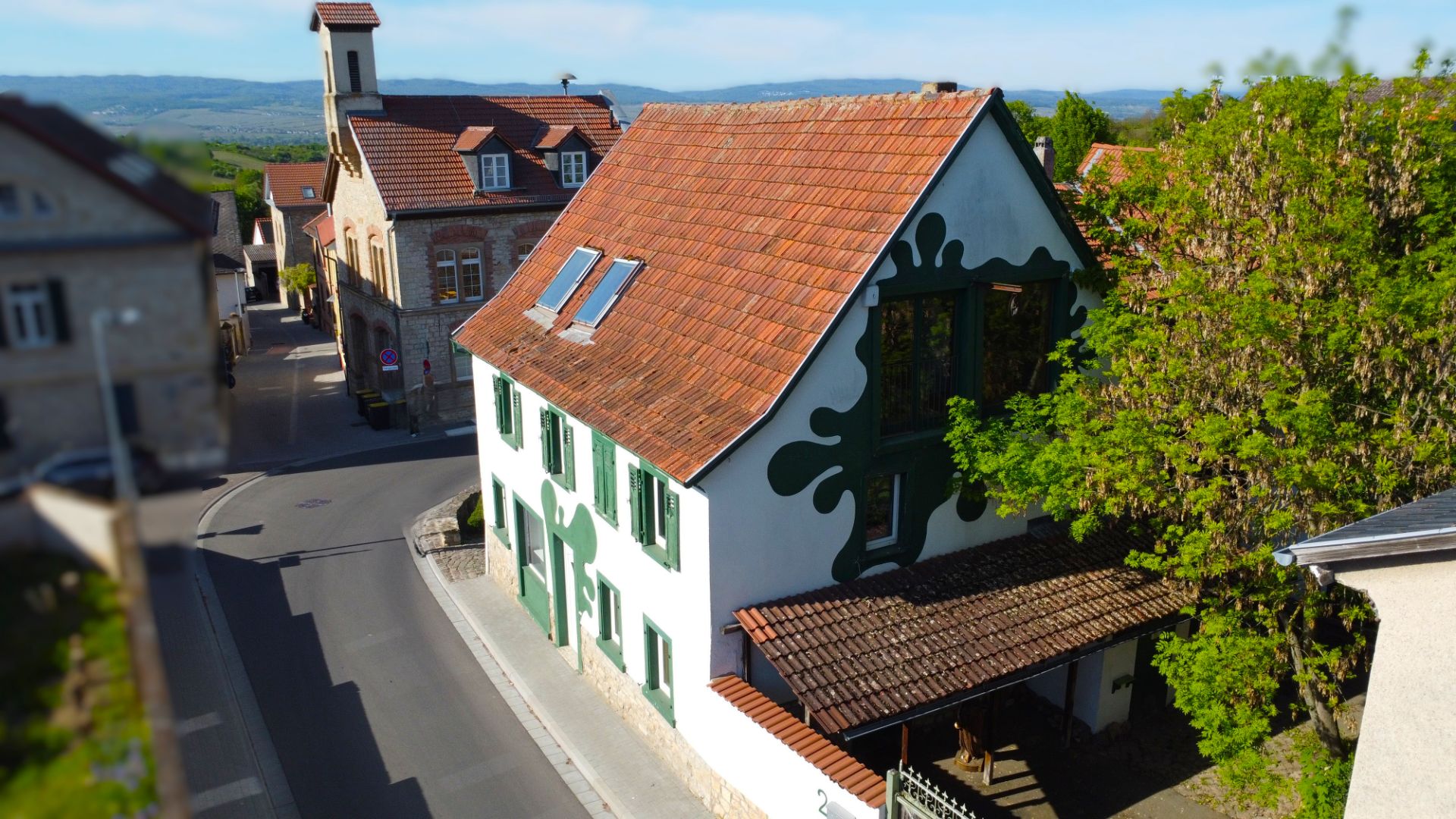 Weißes Wohnhaus mit grünem Ziermuster, rotem Dach und großen Fenstern, an einer Straßenkreuzung gelegen.