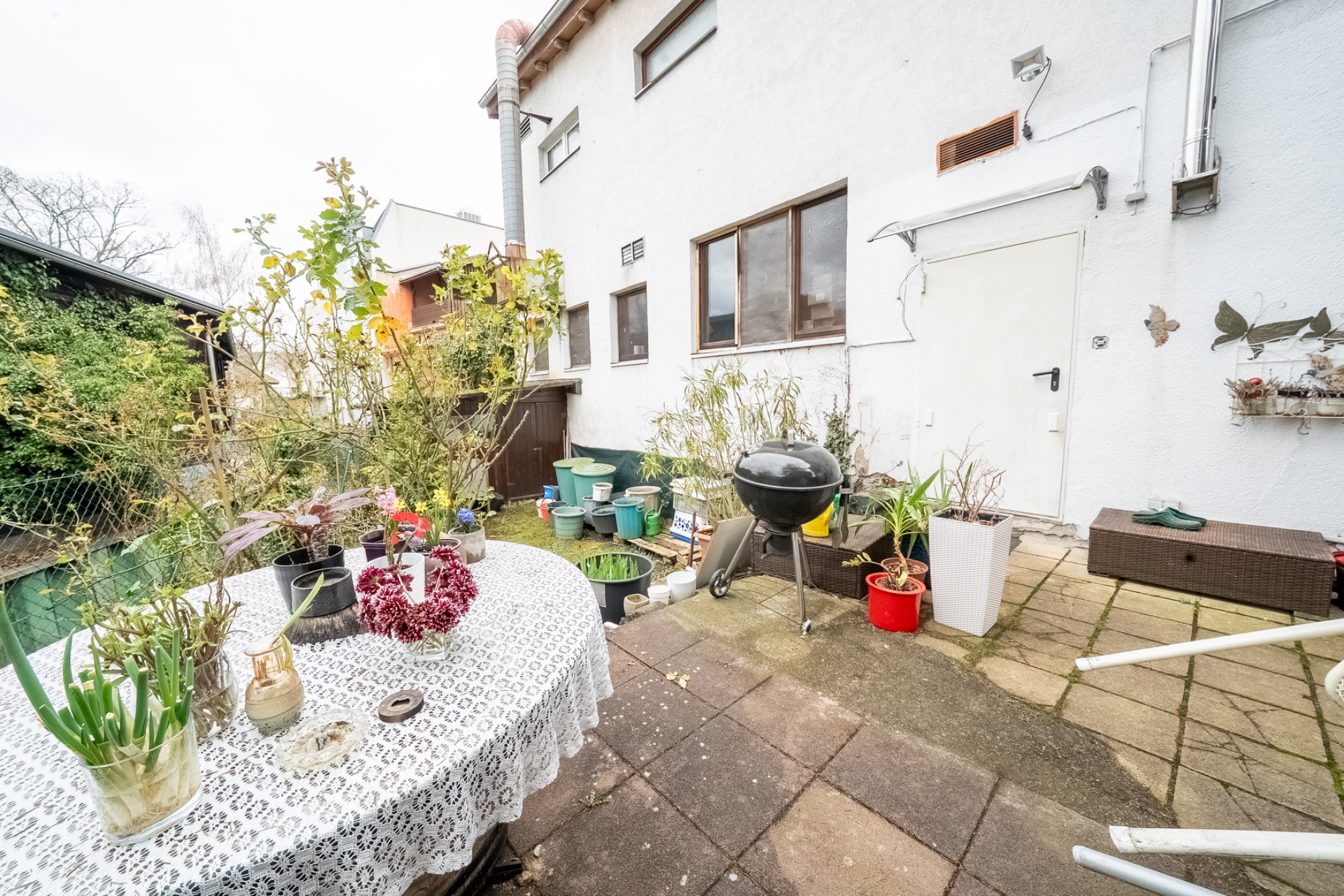 Kleine Terrasse mit Tisch, vielen Pflanzen und einem Grill neben einem weißen Wohnhaus.