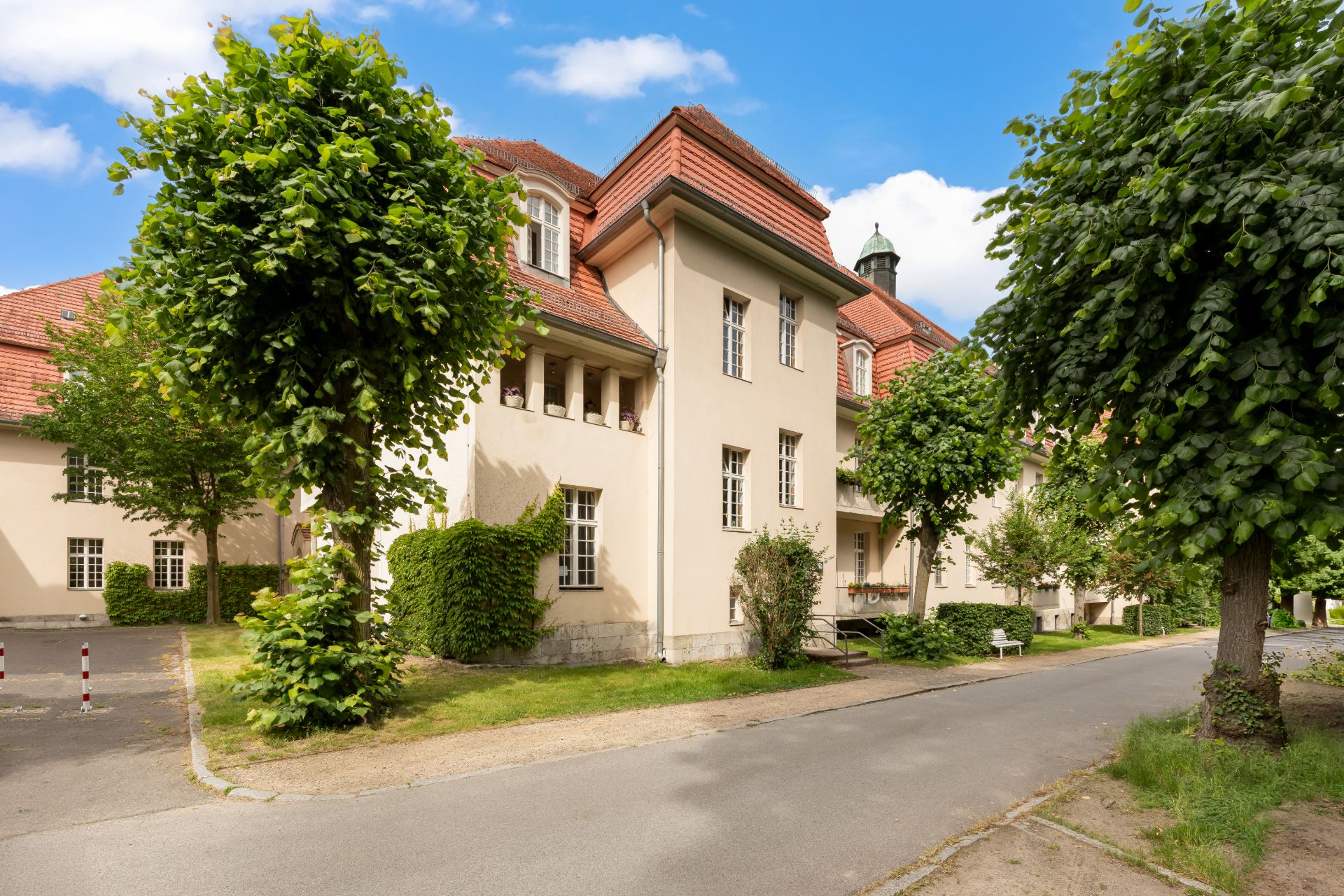 Großes Wohngebäude mit rotem Dach, umgeben von Bäumen und einer Straße im Vordergrund.