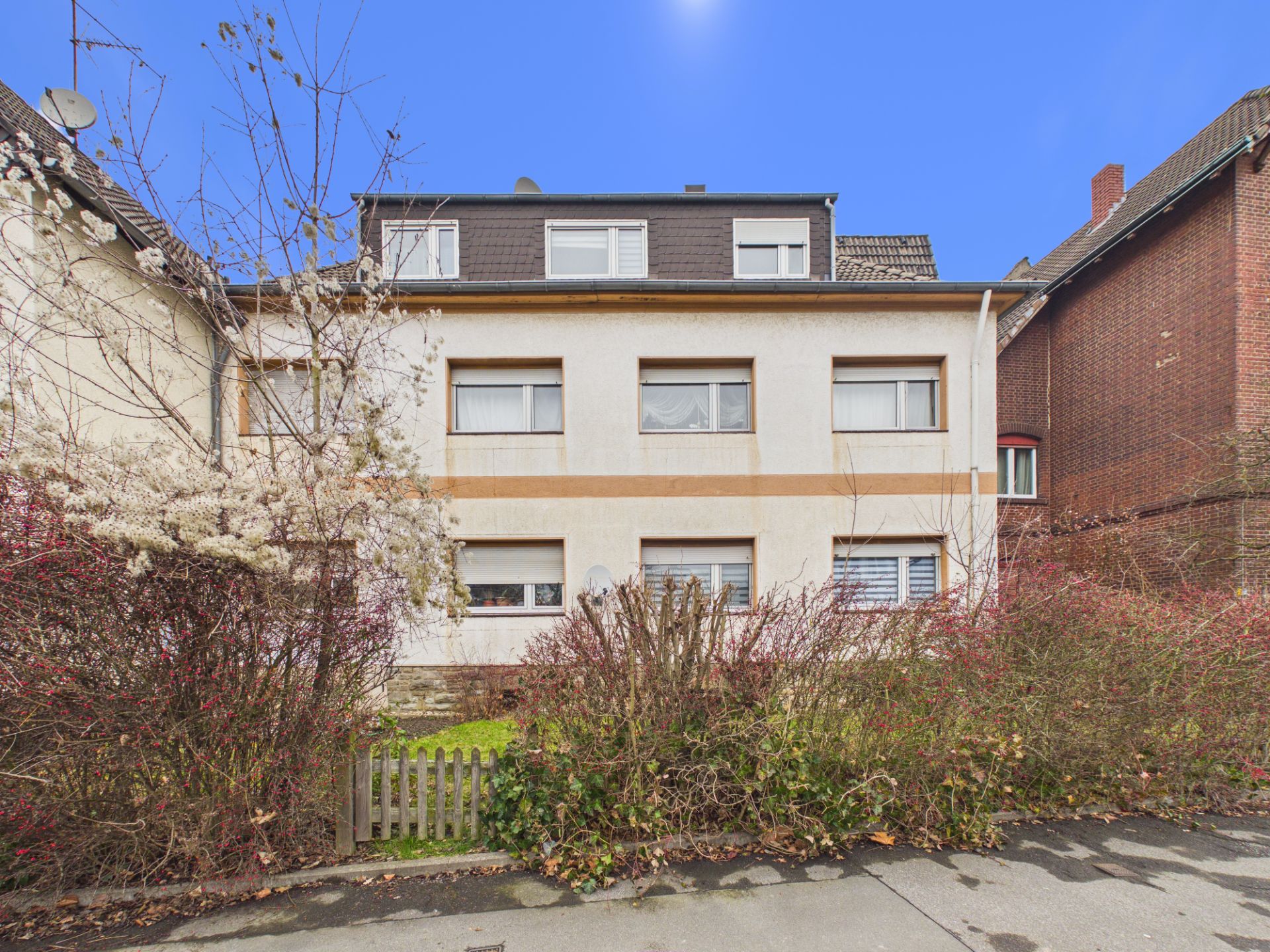 Zweistöckiges Wohnhaus mit Satteldach, mehreren Fenstern und Vorgarten mit Sträuchern.