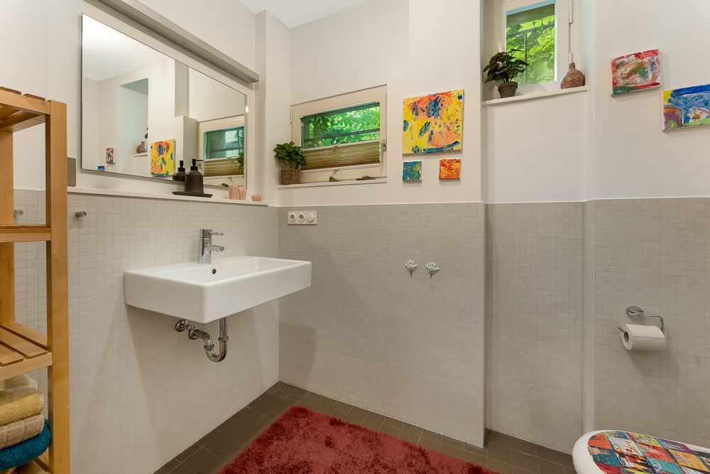 Helles Badezimmer mit Waschbecken, Spiegel, Fenster, bunter Kinderkunst und rotem Teppich.