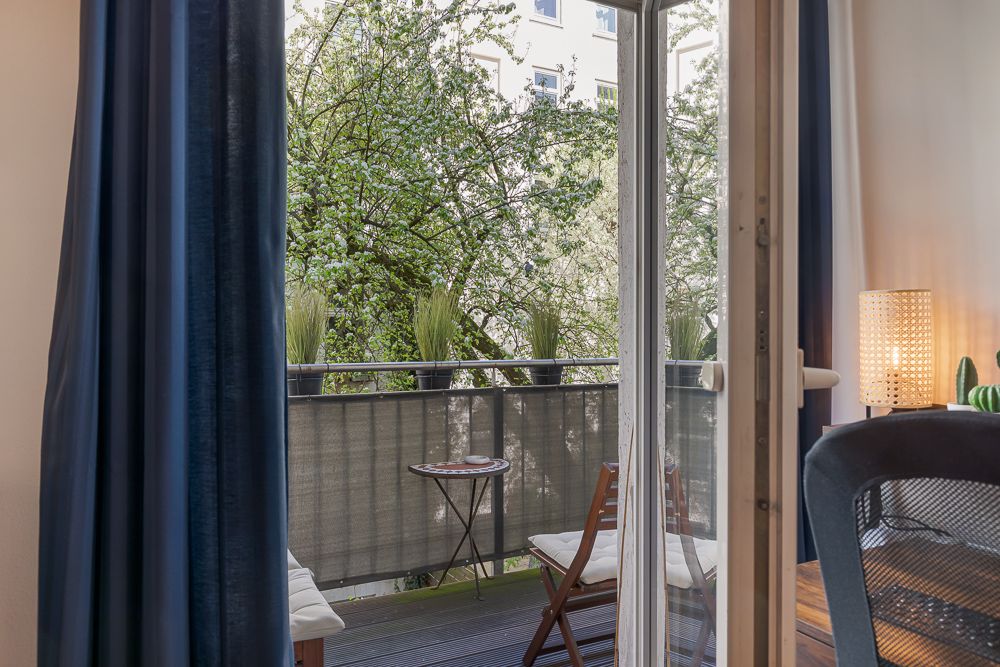 Offene Balkontür mit Blick auf Balkon mit zwei Stühlen, Tisch und Pflanzen, Bäume im Hintergrund.