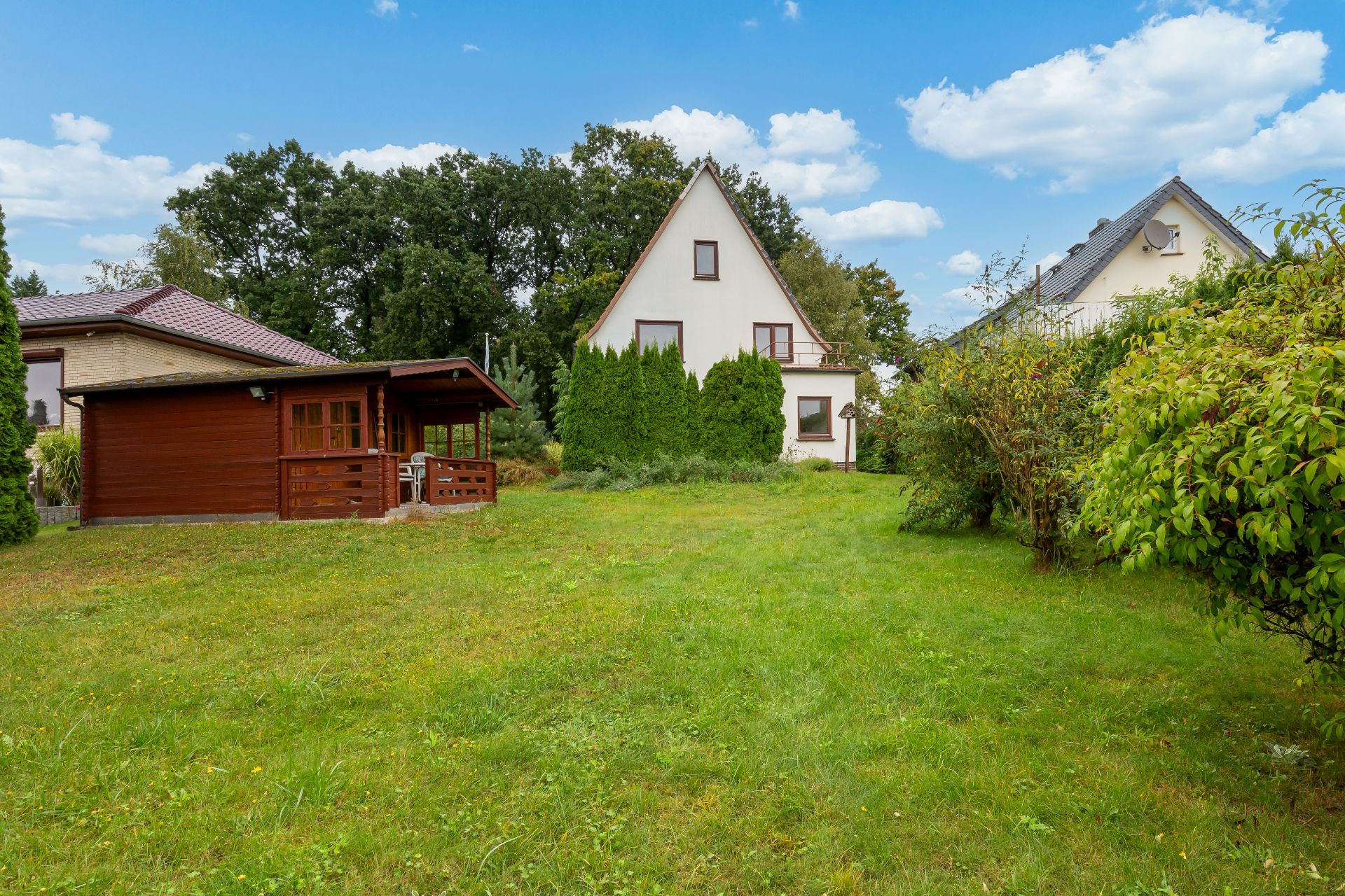 Grüner Garten mit Rasen, Holzlaube und Haus im Hintergrund bei blauem Himmel.
