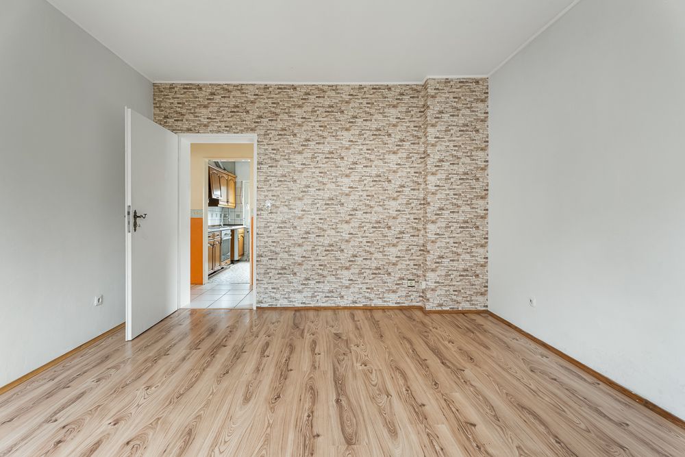 Leeres Zimmer mit Holzfußboden und offener Tür zur Küche, Wand mit Steinoptik-Tapete.