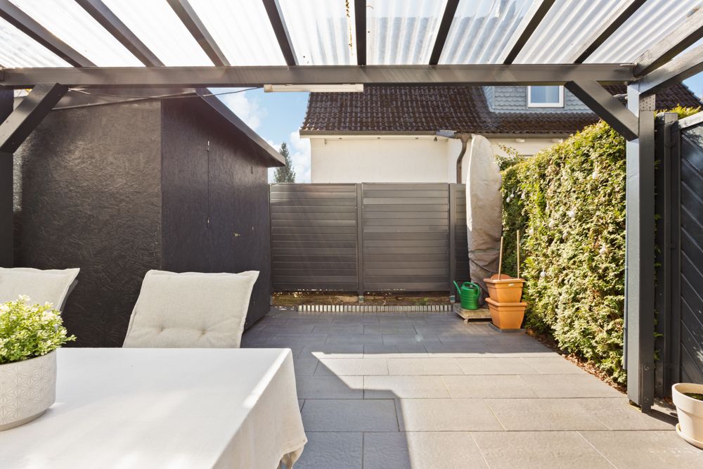 Überdachte Terrasse mit Tisch, Stühlen, Pflanze, Schuppen und Hecke an der Seite.