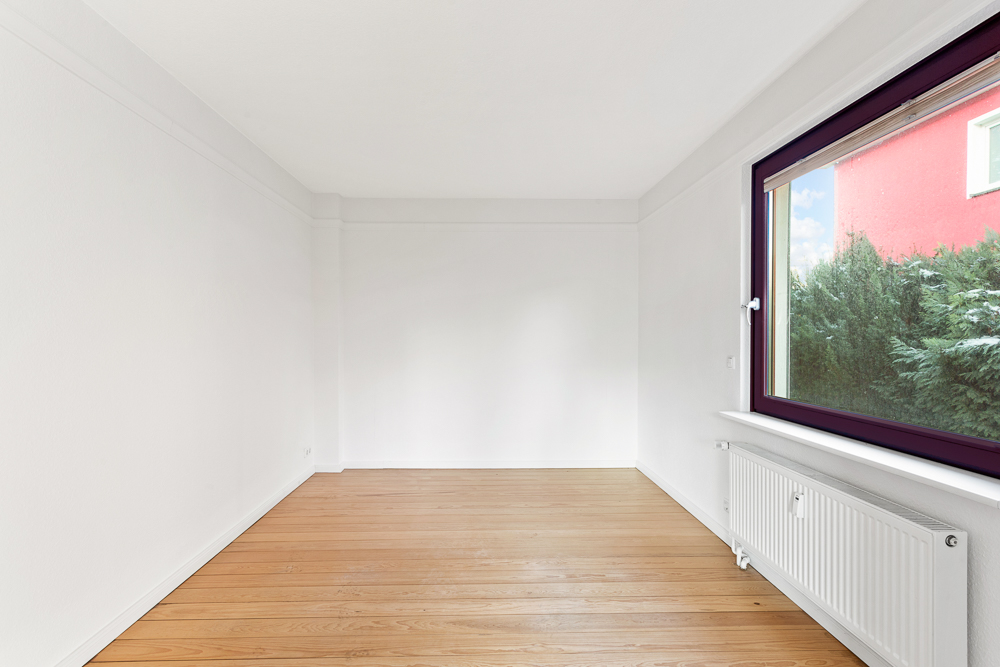 Leerer Raum mit weiß gestrichenen Wänden, Holzboden und großem Fenster mit Blick nach draußen.