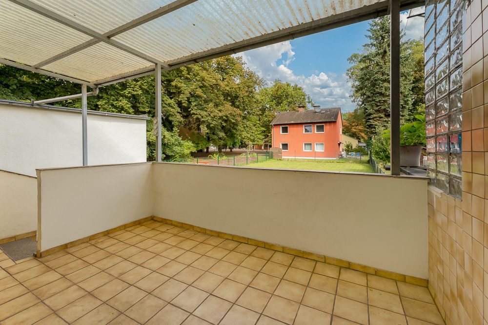 Überdachte Terrasse mit Fliesenboden und Blick auf Garten und rotes Wohnhaus.