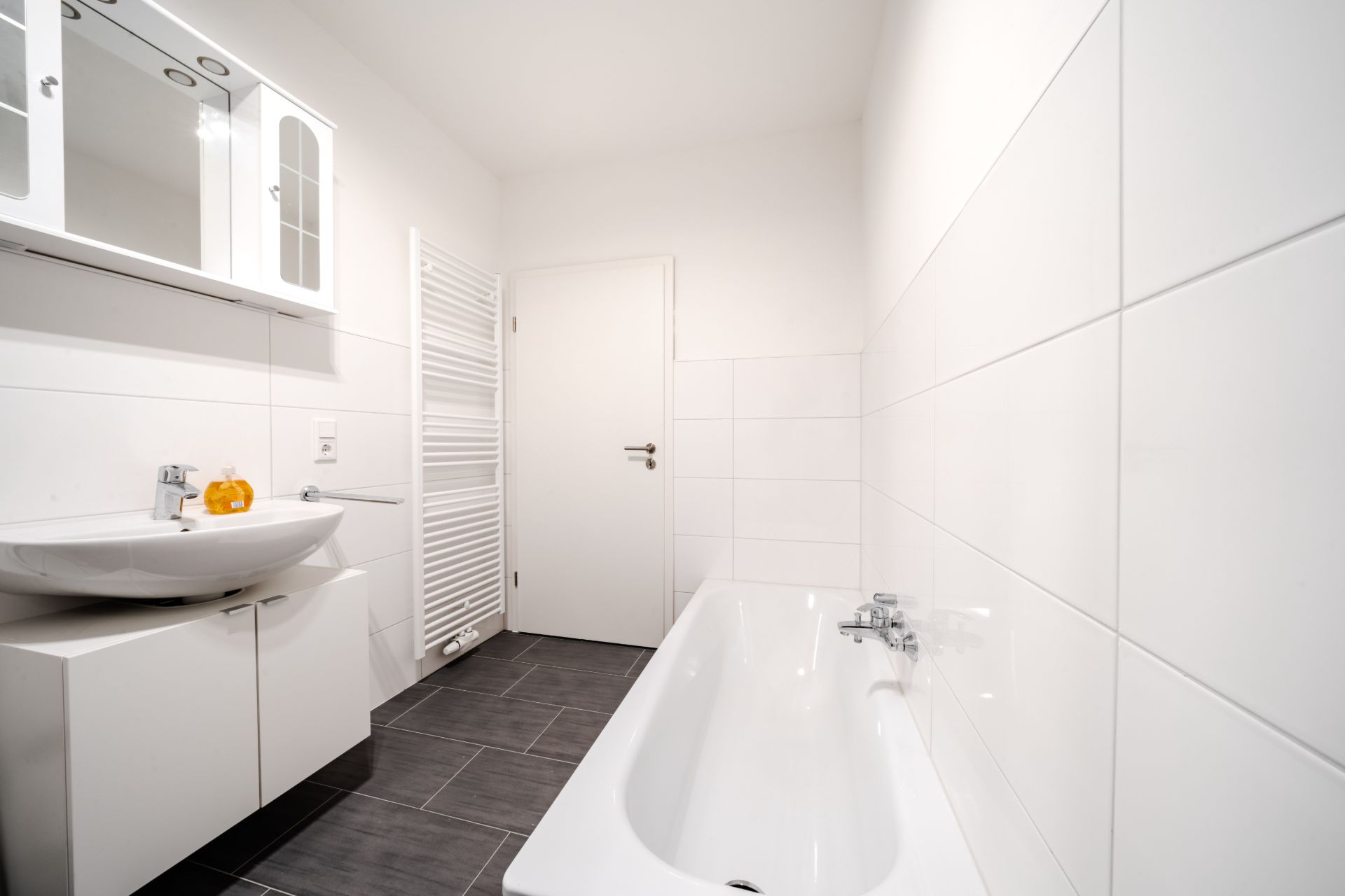 Helles Badezimmer mit Badewanne, Waschbecken, Spiegel und Handtuchheizkörper.