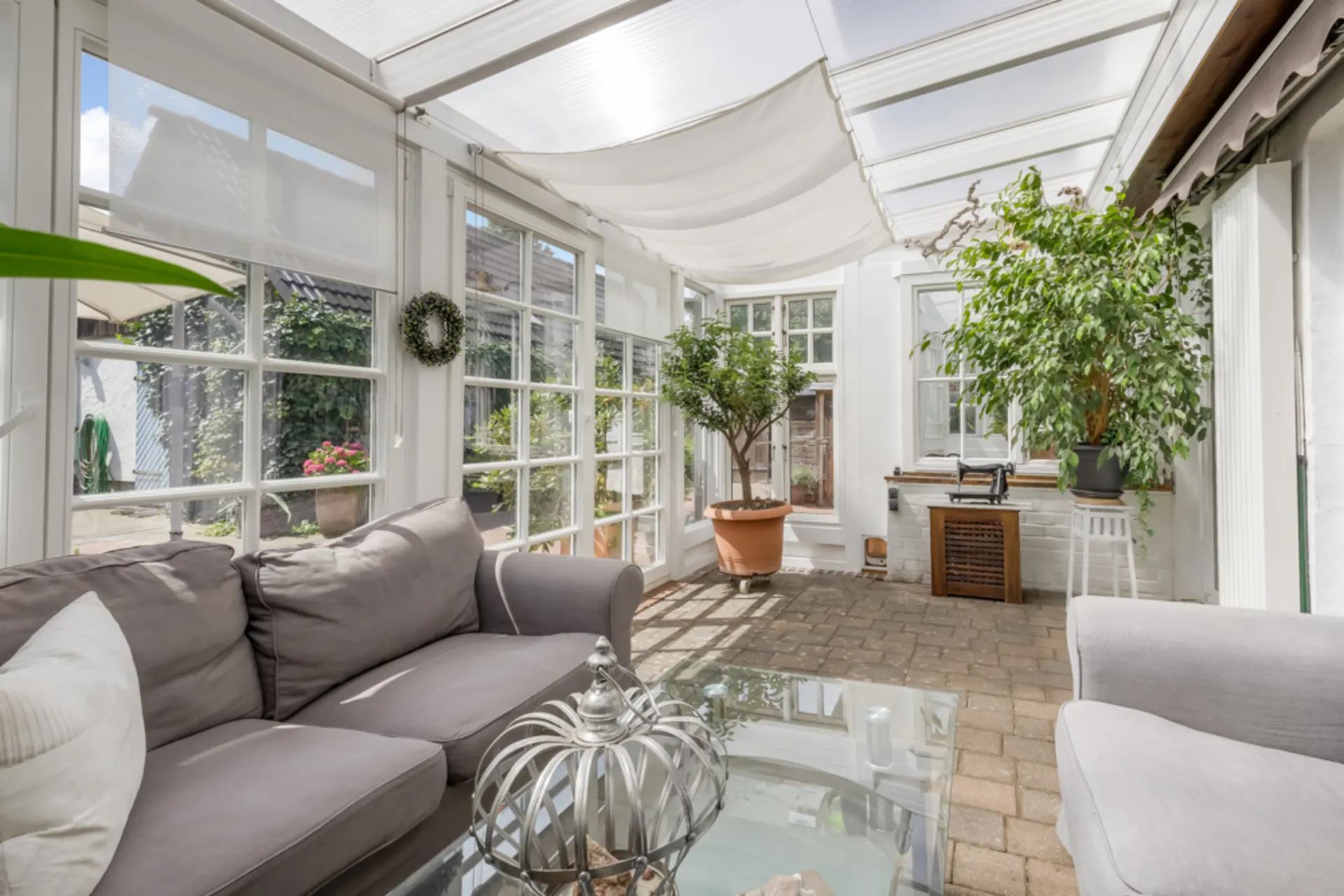 Heller Wintergarten mit grauen Sofas, Glastisch und großen Topfpflanzen.