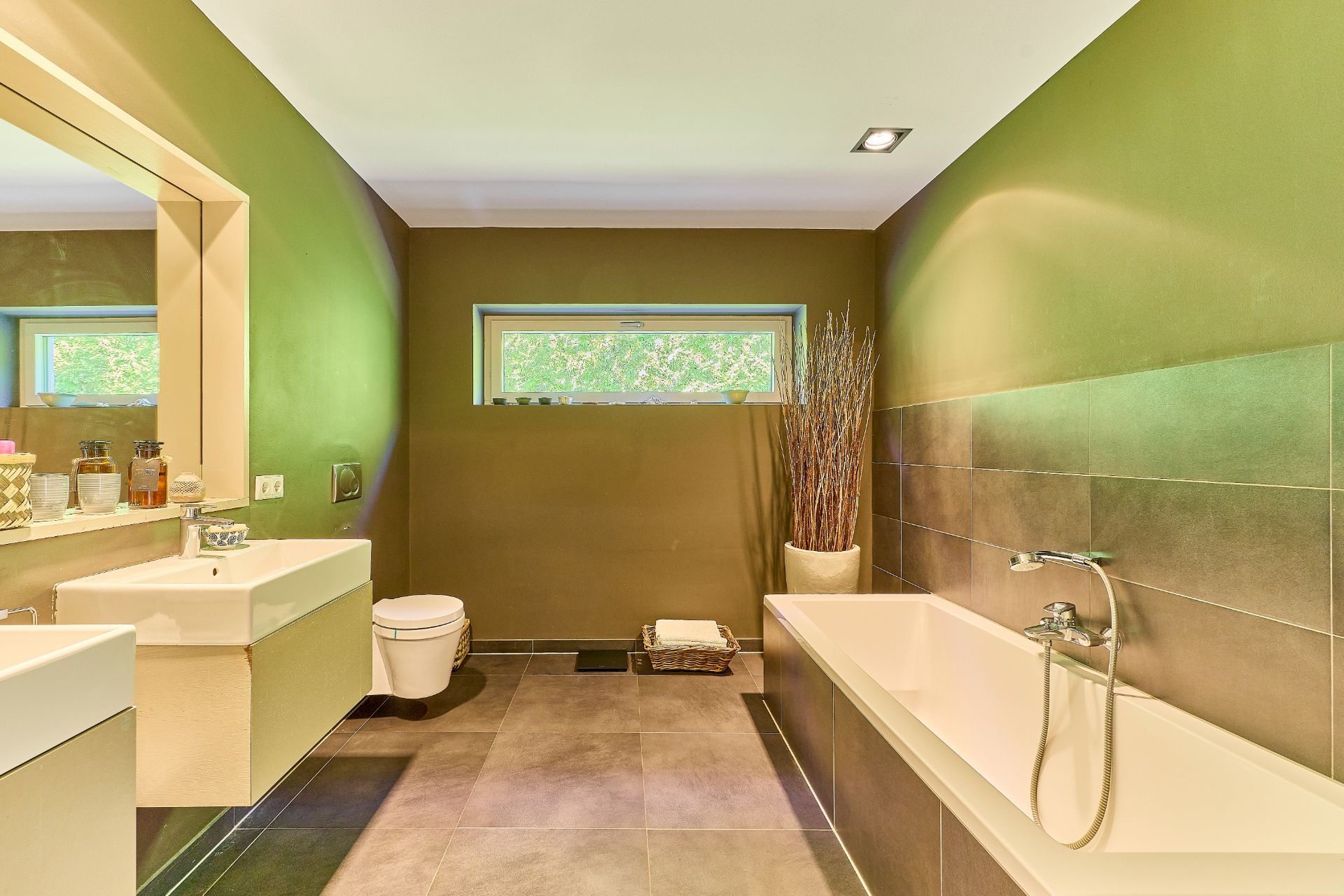 Modernes Badezimmer mit Badewanne, Waschbecken, WC und großem Spiegel, grünen Wänden und Fenster.