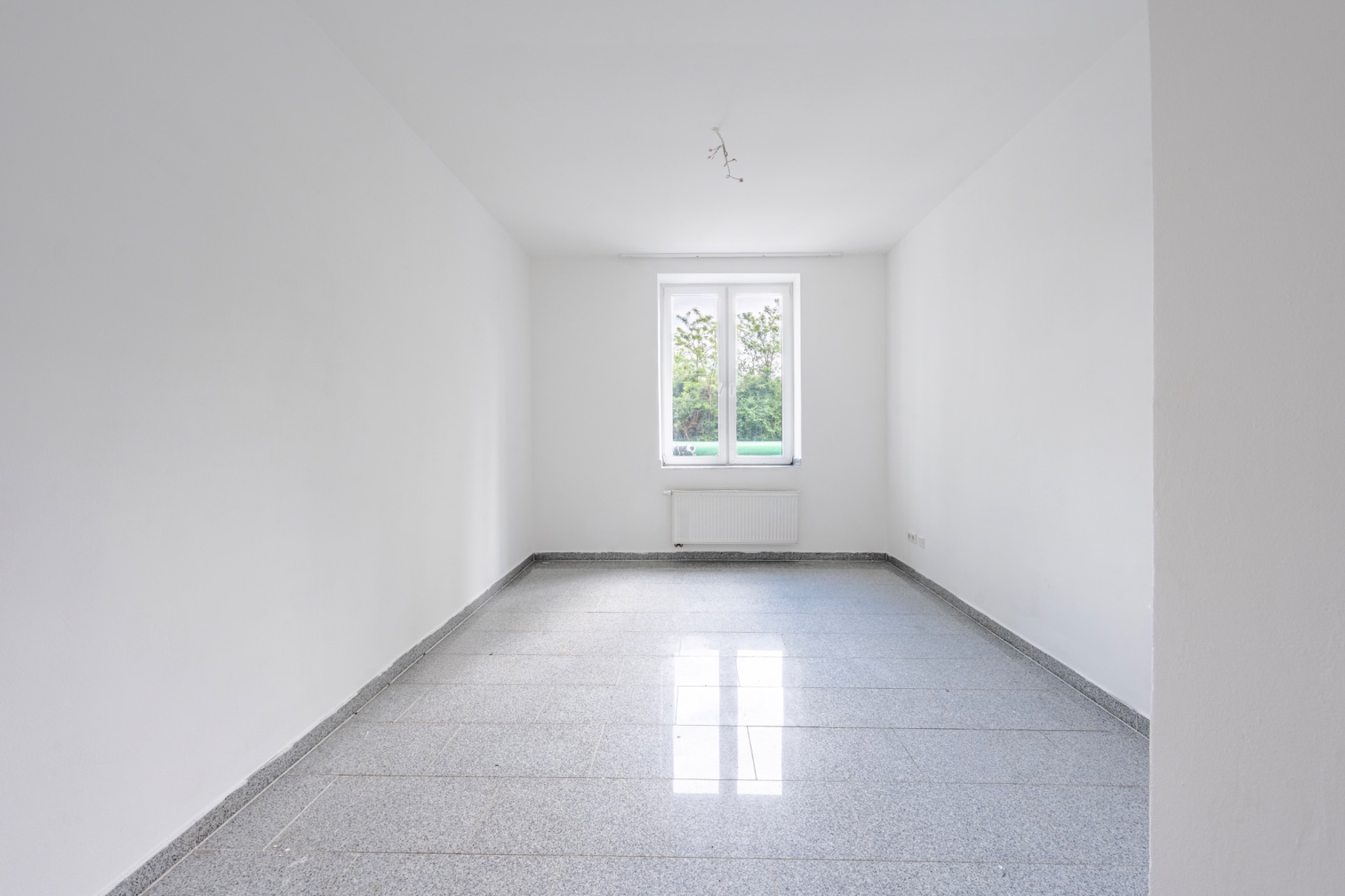 Leerer, heller Raum mit weißen Wänden, Fliesenboden und einem Fenster am Ende.