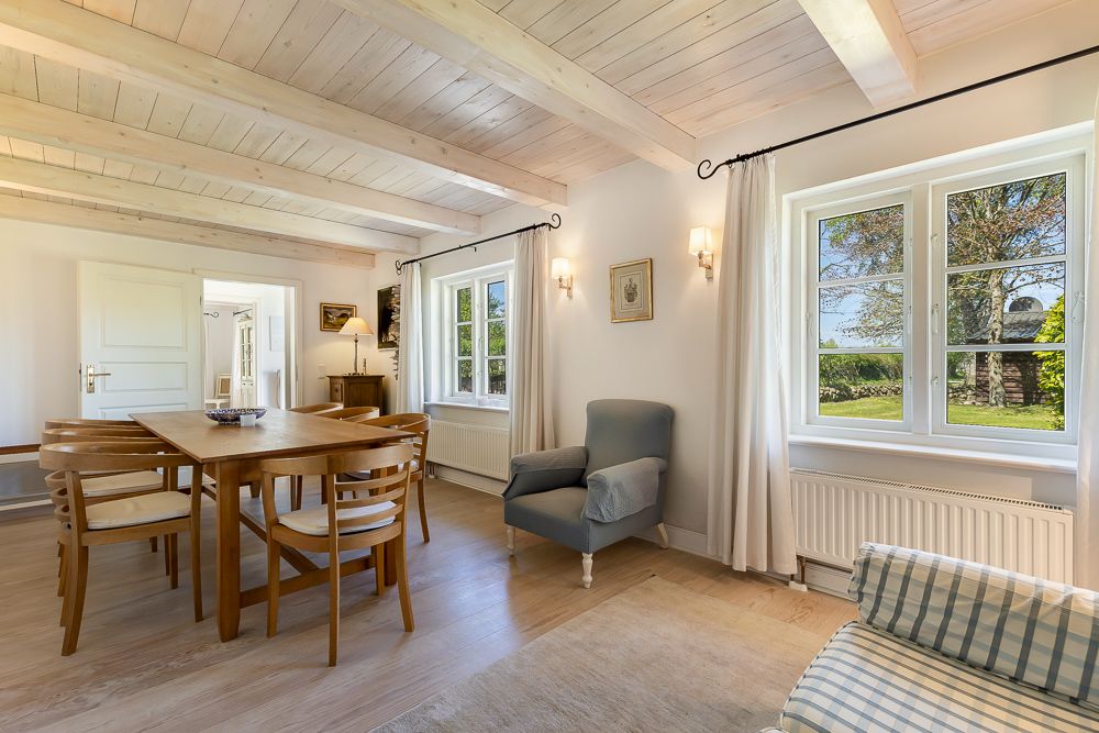 Helles Esszimmer mit Holztisch, sechs Stühlen, Sessel und großen Fenstern mit Blick nach draußen.