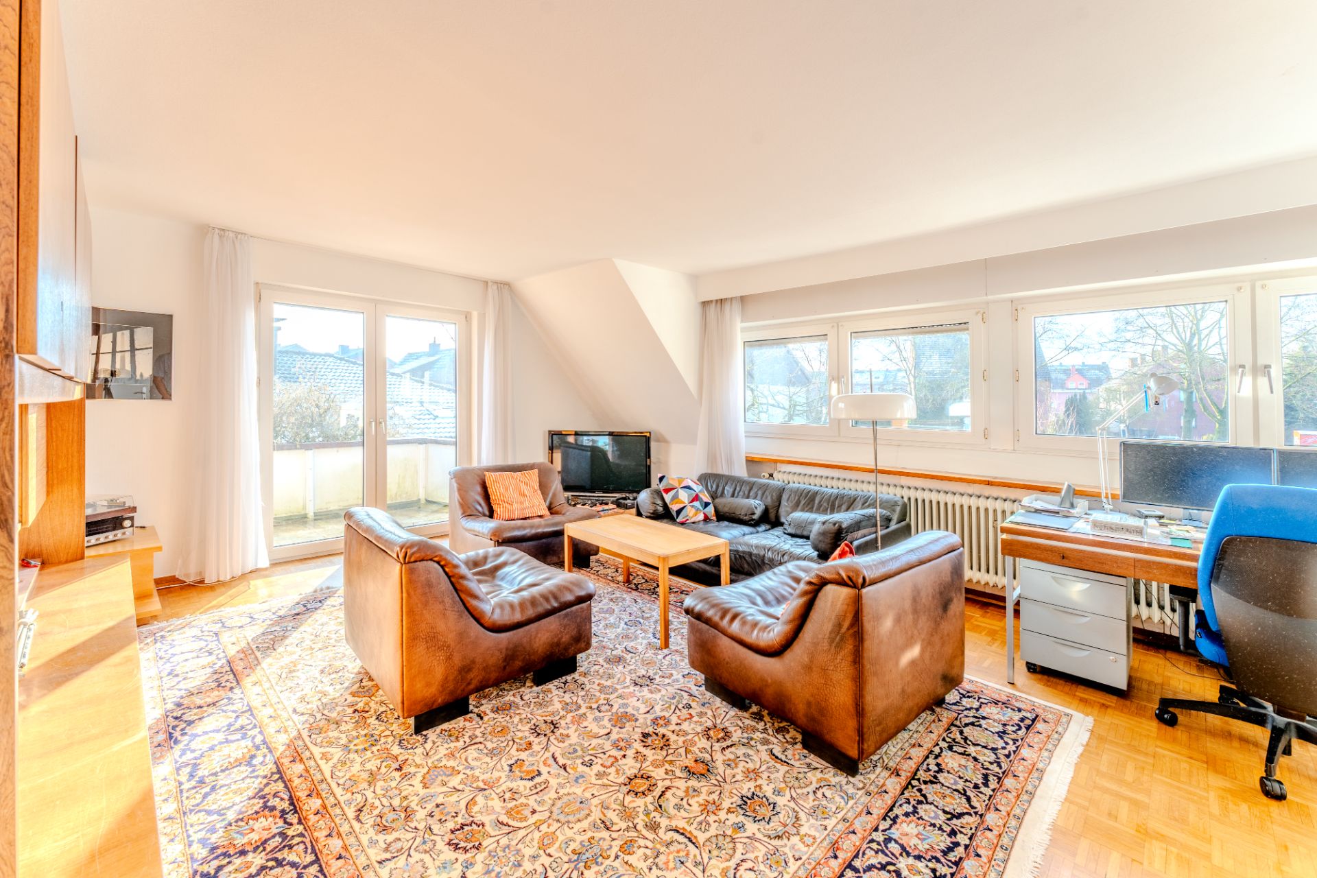 Helles Wohnzimmer mit Ledersesseln, Sofa, Teppich, TV, großem Fenster und Schreibtisch mit Stuhl.