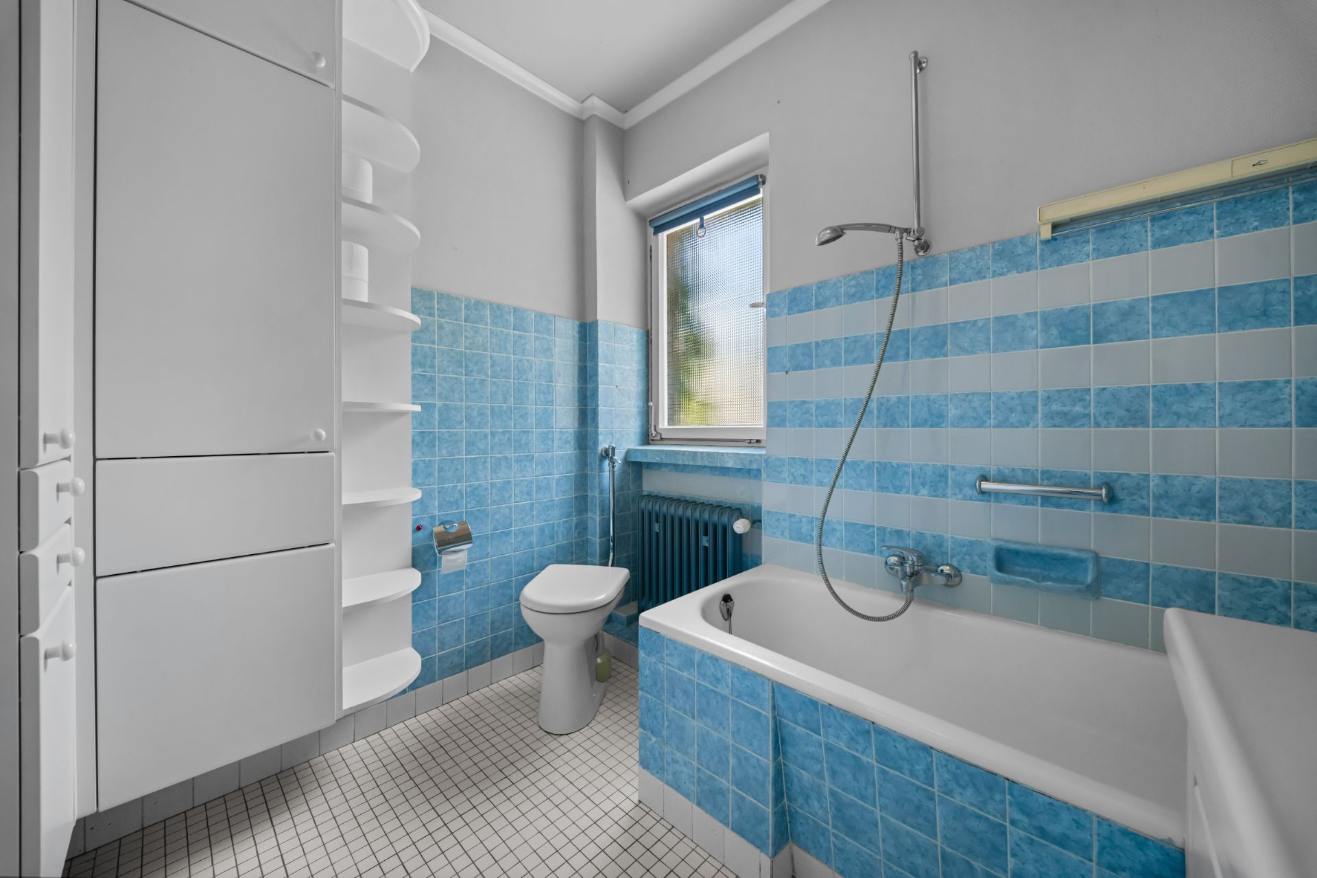 Helles Badezimmer mit blauen Fliesen, Badewanne, Toilette, Fenster und weißen Schränken.