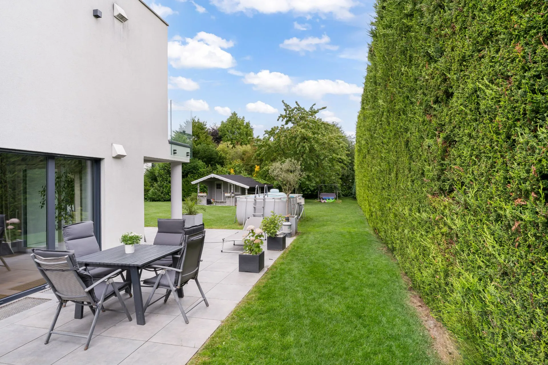 Terrasse mit Gartentisch und Stühlen neben Rasen, hohe Hecke und Gartenhaus im Hintergrund.