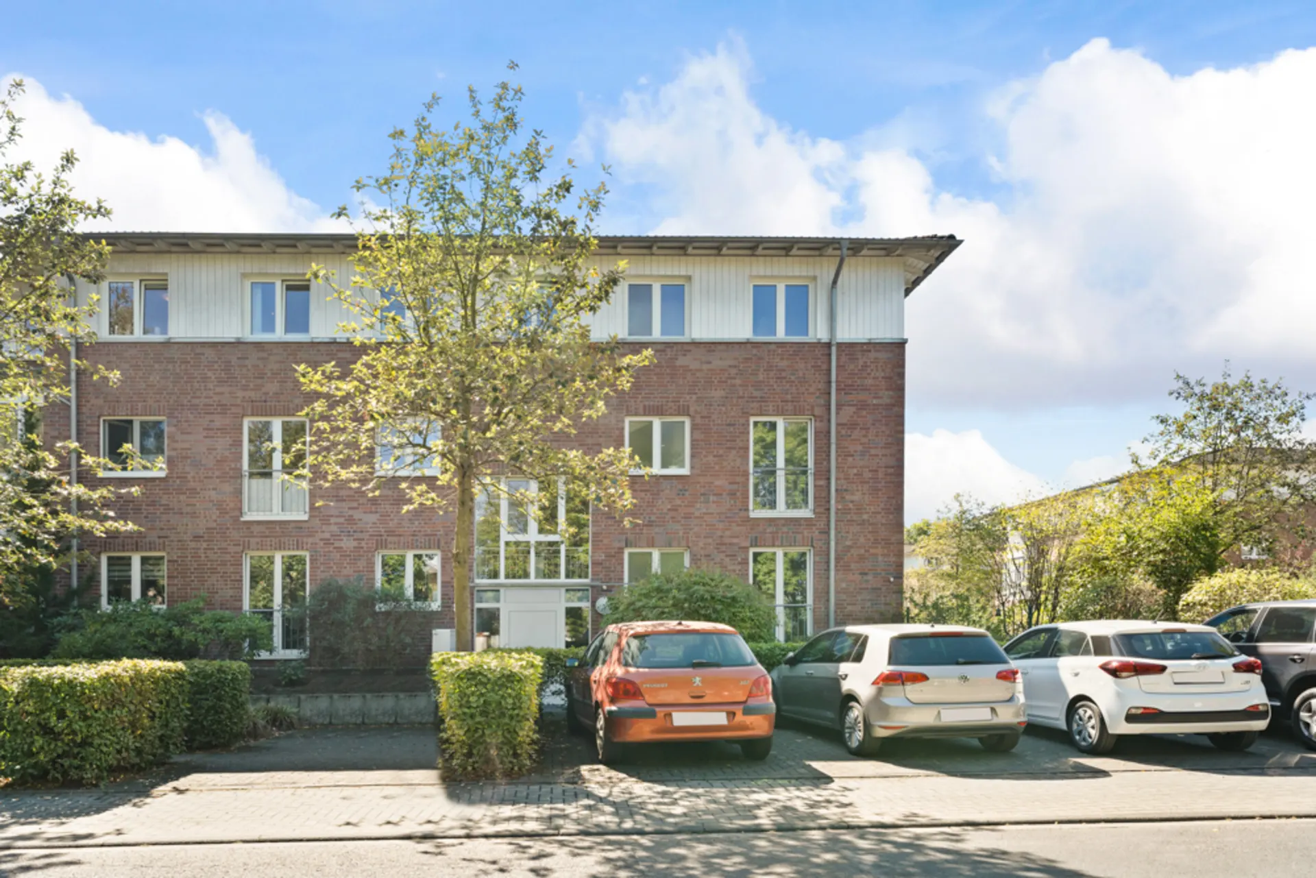 Dreistöckiges Wohnhaus aus rotem Backstein mit parkenden Autos und Bäumen davor.