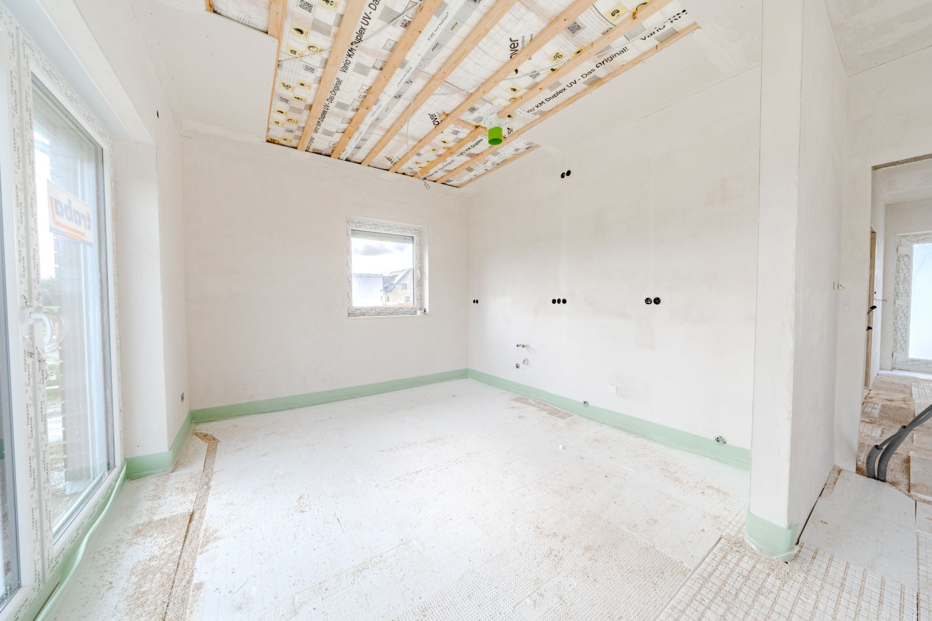 Leerer Raum im Bau mit sichtbarer Decke, Fenster und Glastür, Wände und Boden noch unfertig.