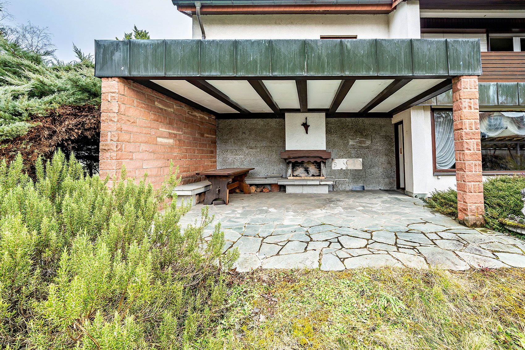 Überdachte Terrasse mit gemauertem Grill, Holzbänken und Natursteinboden, umgeben von Sträuchern.