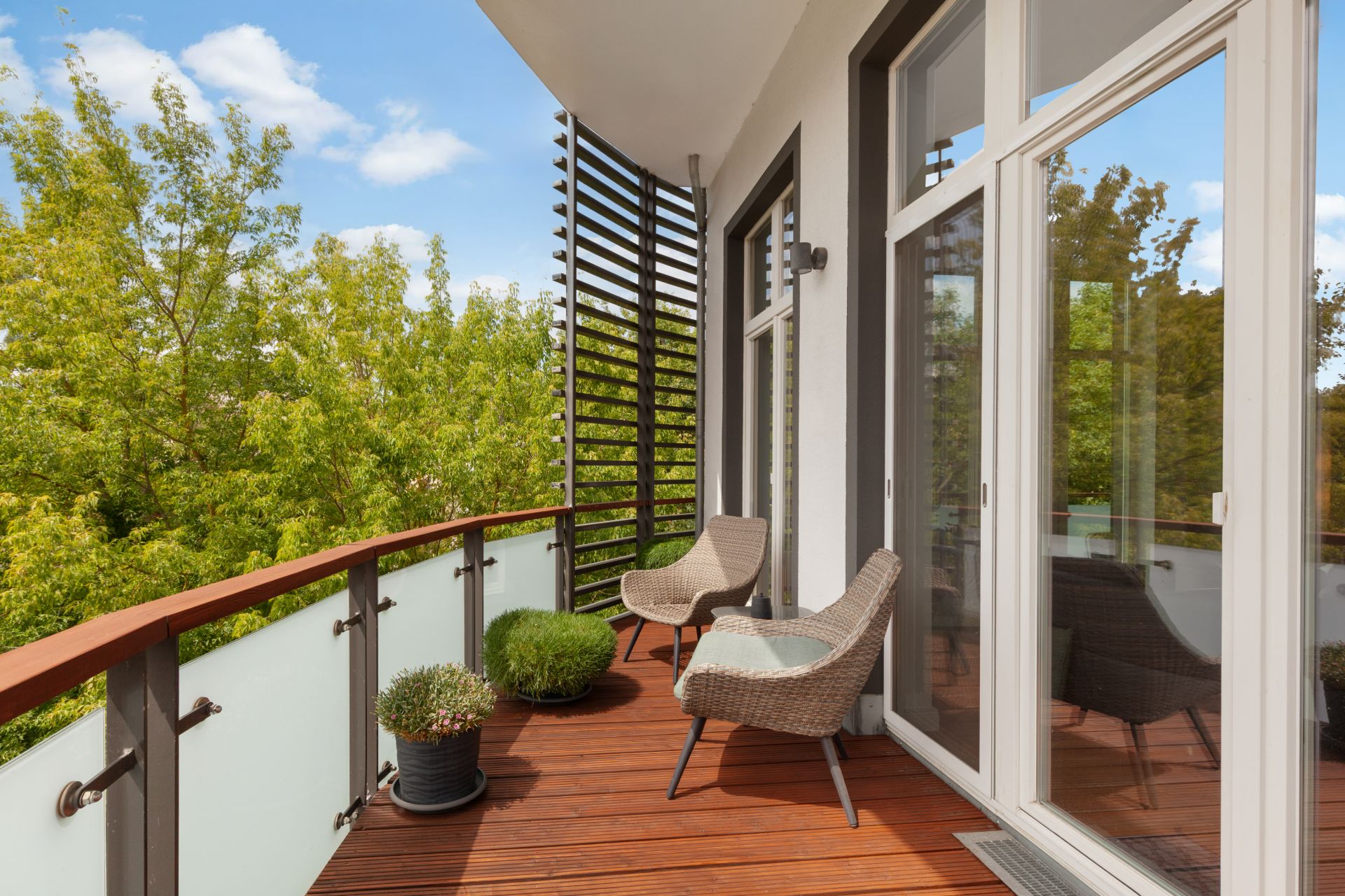 Balkon mit zwei Stühlen, Pflanzen und Glasgeländer, umgeben von Bäumen.