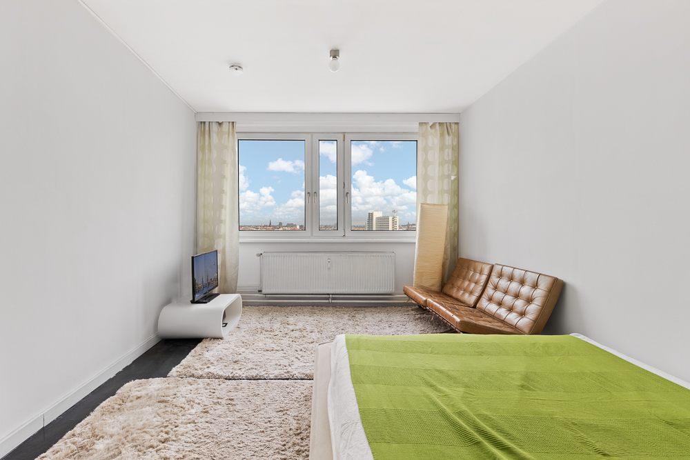 Helles Zimmer mit Fenster, braunem Sofa, Teppich, TV und Bett mit grüner Decke.