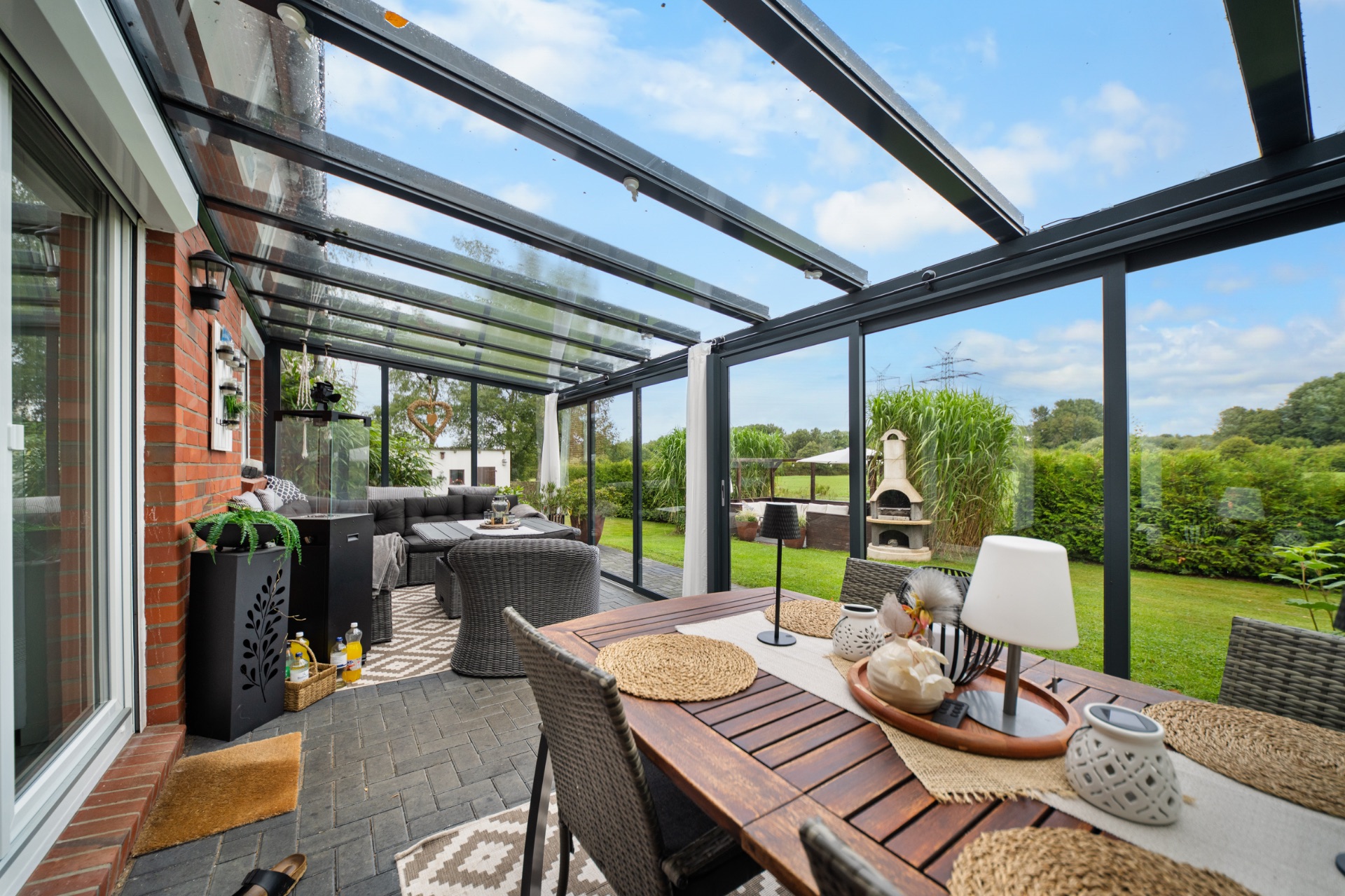 Wintergarten mit Esstisch, Stühlen und Sofa, Blick in einen Garten mit viel Grün.