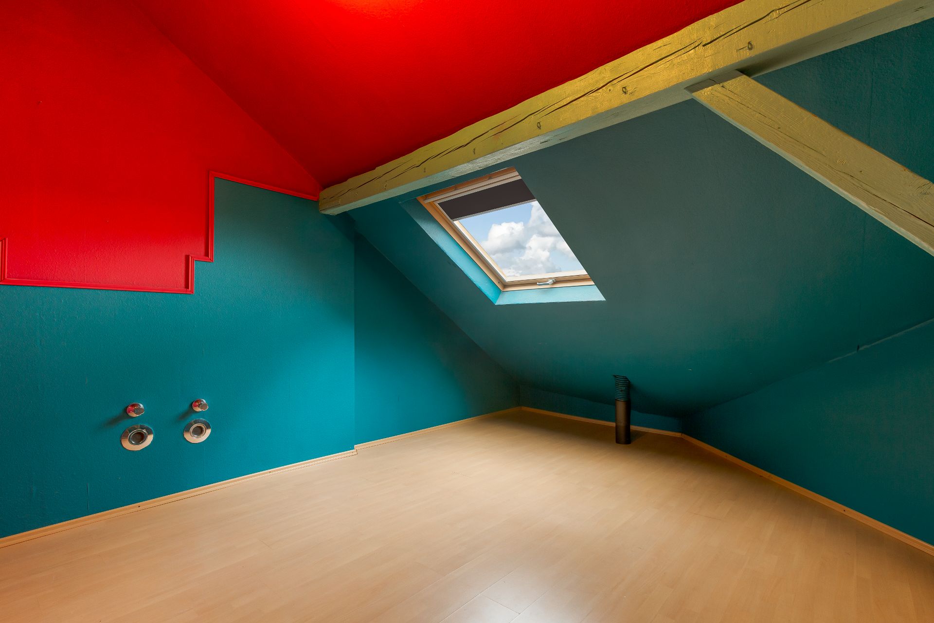 Dachzimmer mit schrägem Fenster, roten und blauen Wänden sowie hellem Holzboden.