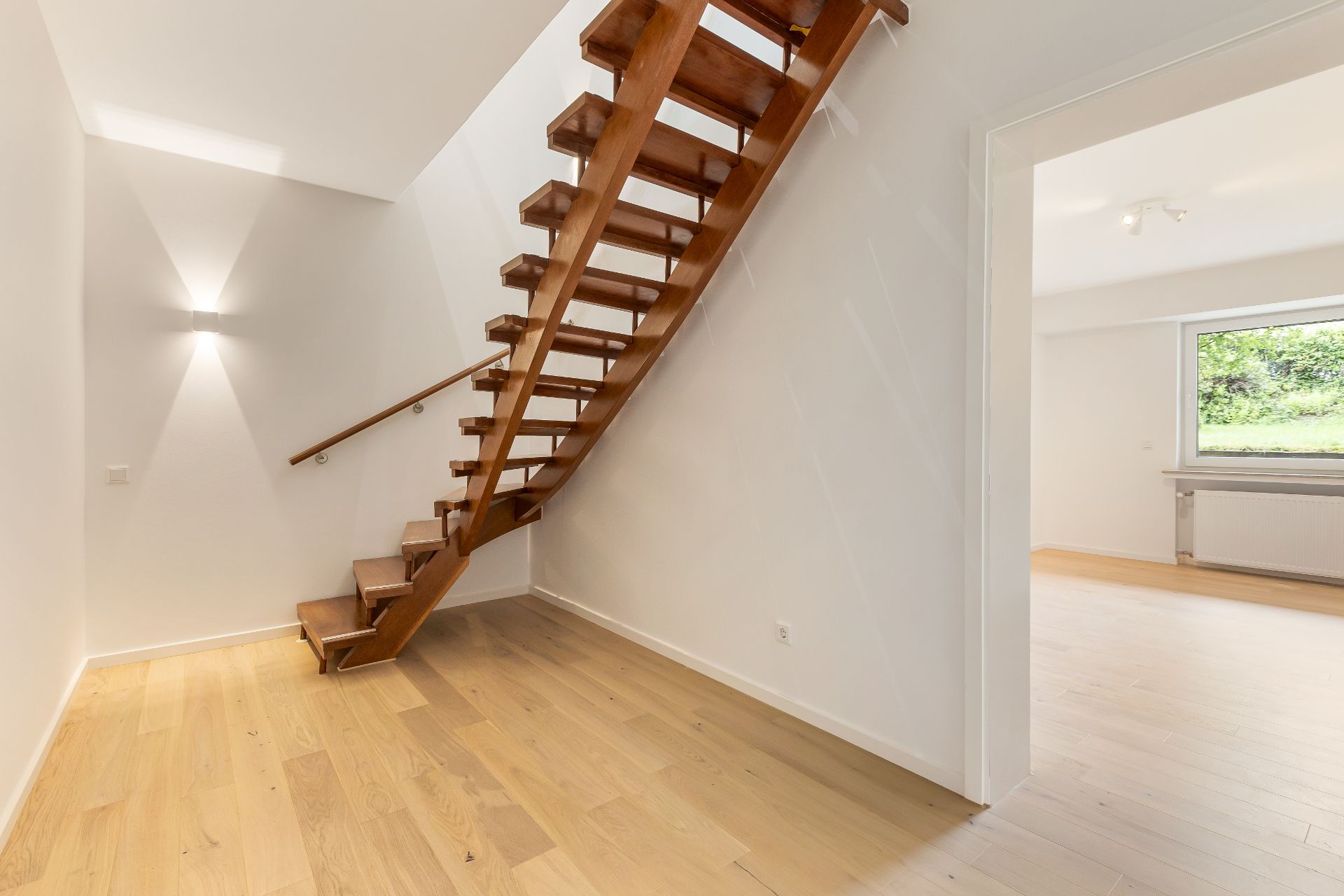 Heller Flur mit Holztreppe und Blick in ein Zimmer mit großem Fenster.