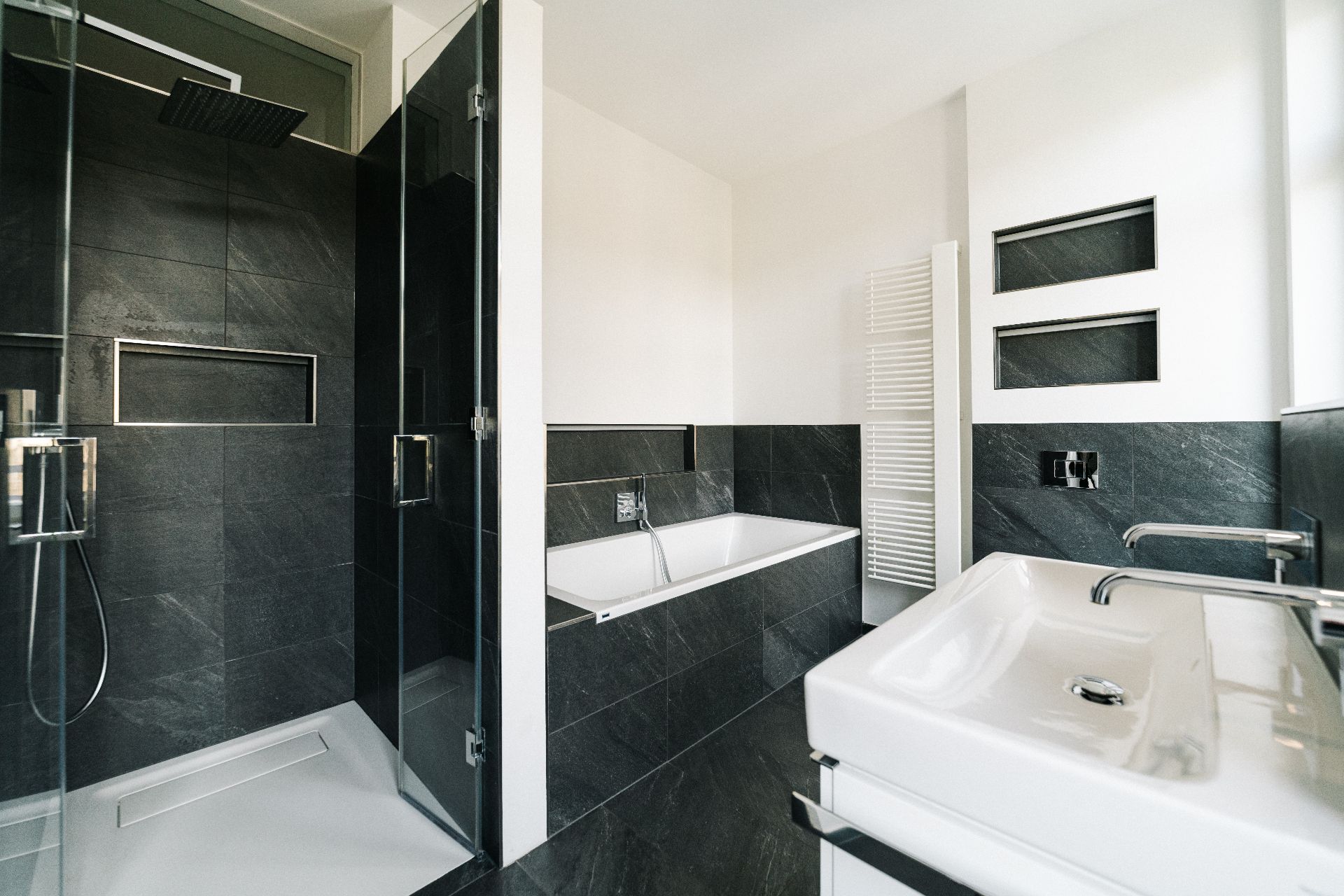 Modernes Badezimmer mit Dusche, Badewanne, Waschbecken und dunklen Fliesen.